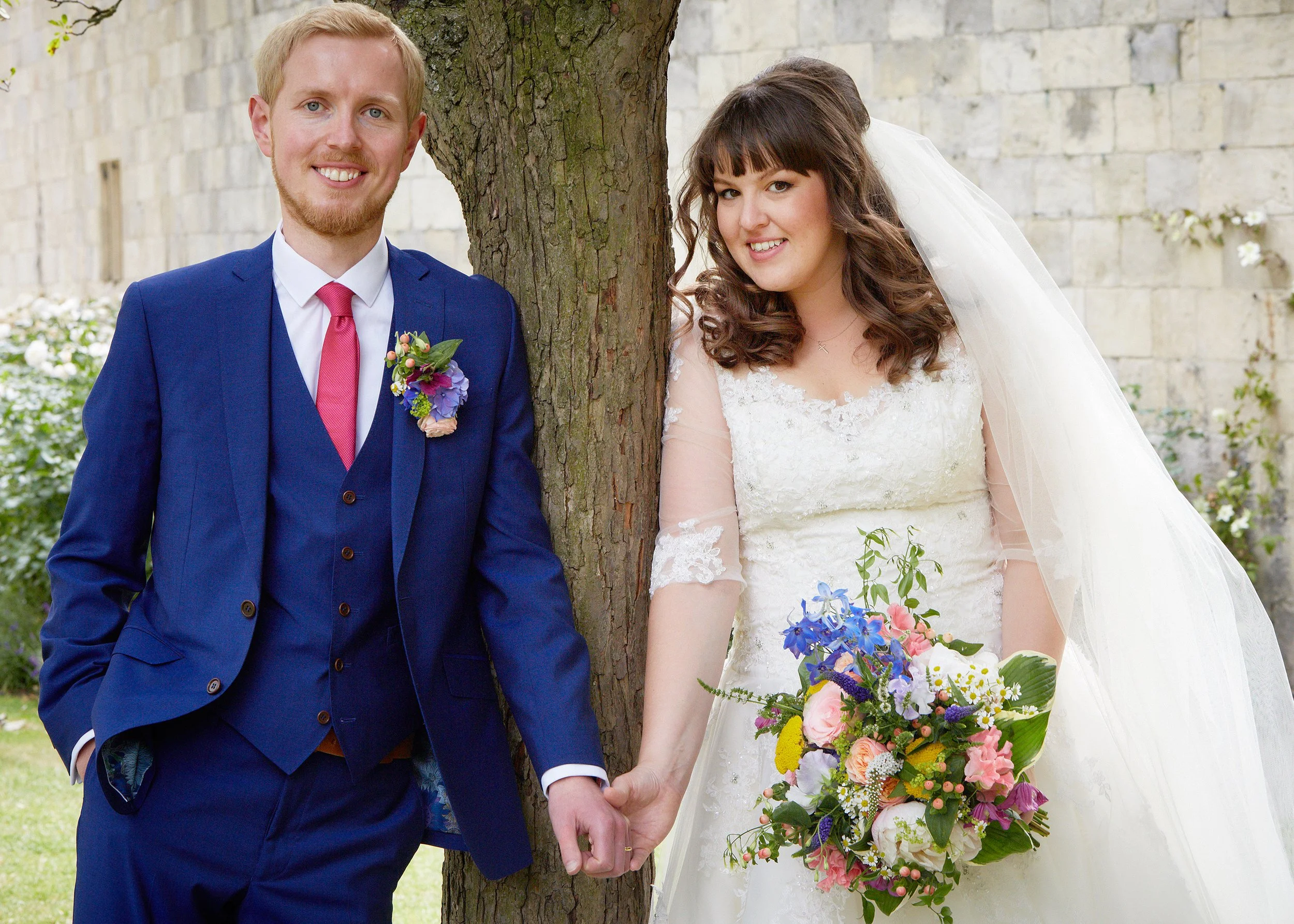 A bride and groom holding hands outdoors near a tree, smiling. The groom is dressed in a blue suit with a pink tie and a floral boutonniere. The bride is wearing a white lace wedding gown and veil, holding a colorful bouquet.