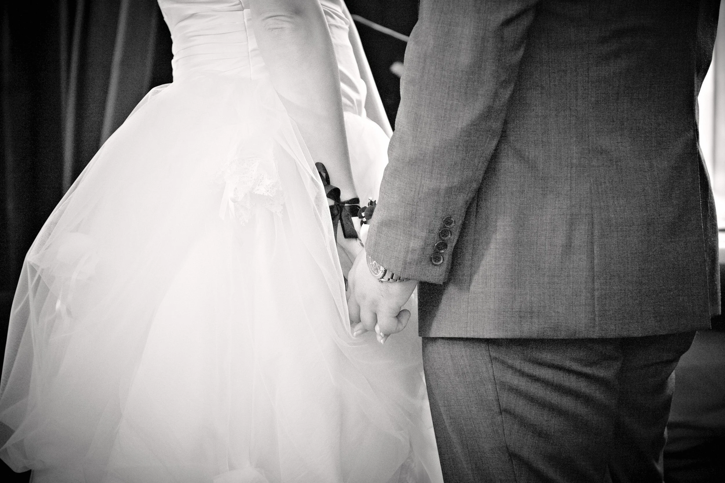 A close-up black and white photo of a couple holding hands during a wedding ceremony, with the bride wearing a white gown and the groom in a gray suit.
