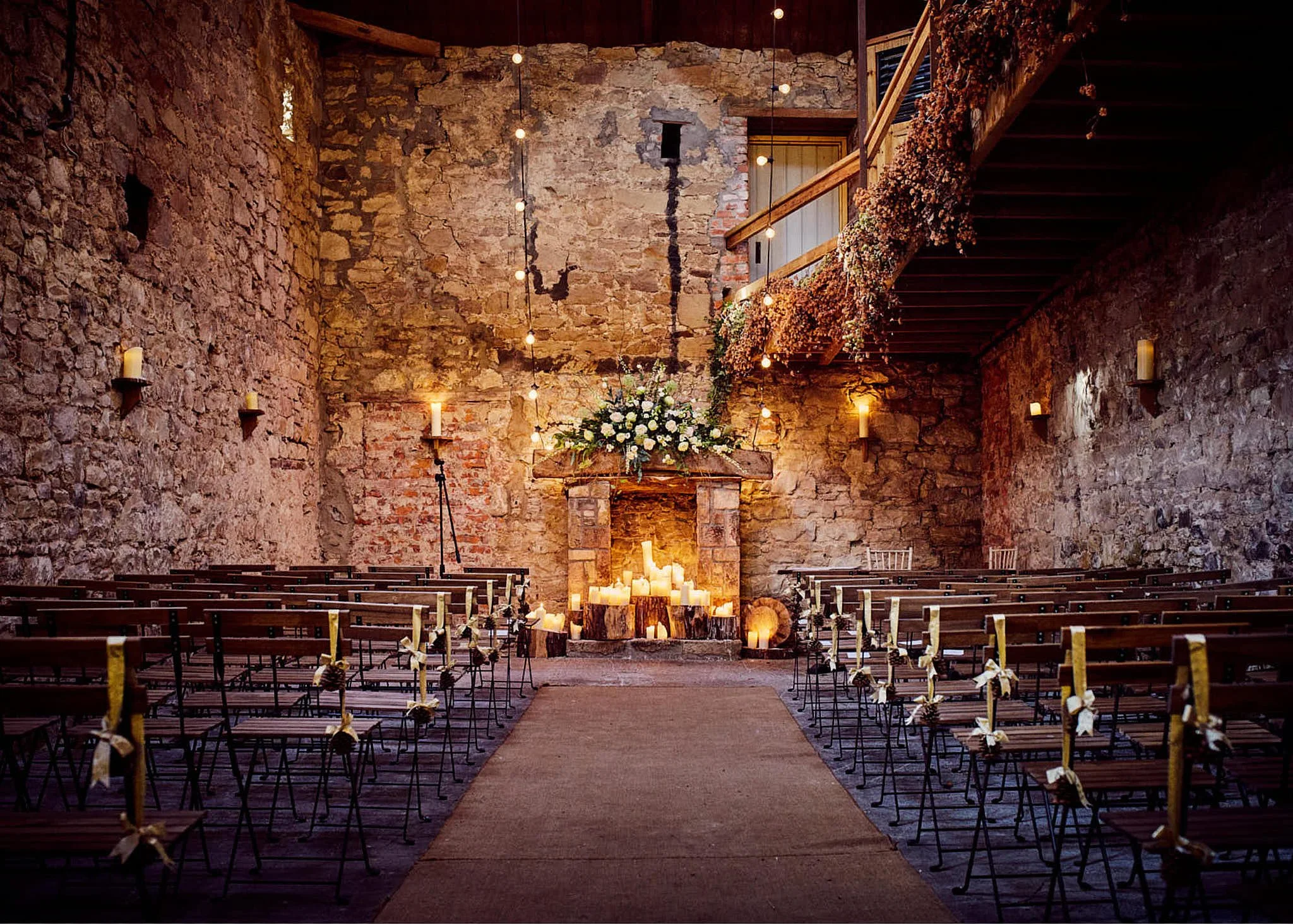 Interior of a rustic wedding venue with stone walls, wooden accents, hanging string lights, candles on the altar, a floral arrangement, and rows of chairs decorated with ribbons.