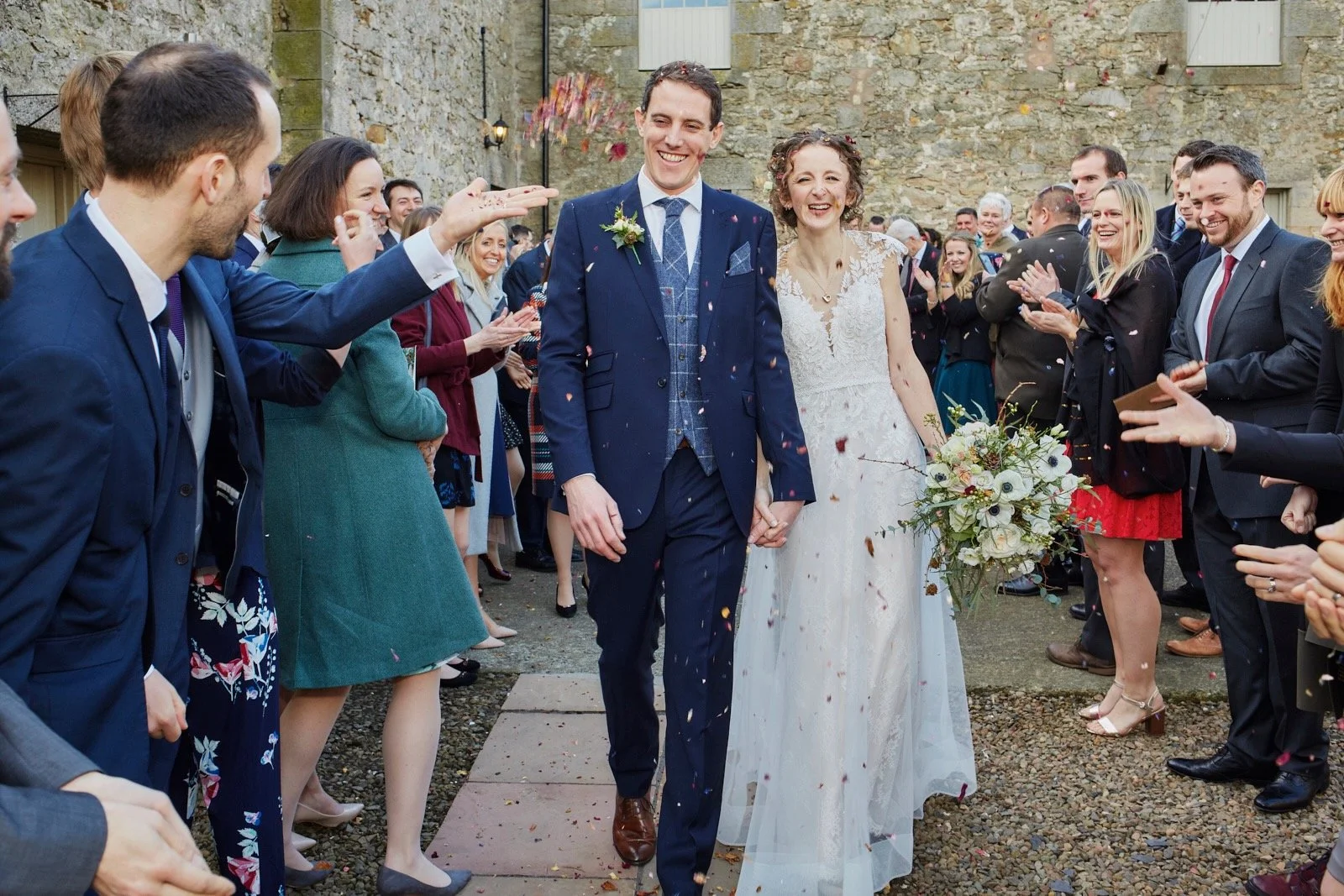 A newlywed couple walking hand in hand through a crowd of wedding guests, with the bride holding a bouquet of flowers and everyone celebrating and throwing confetti.