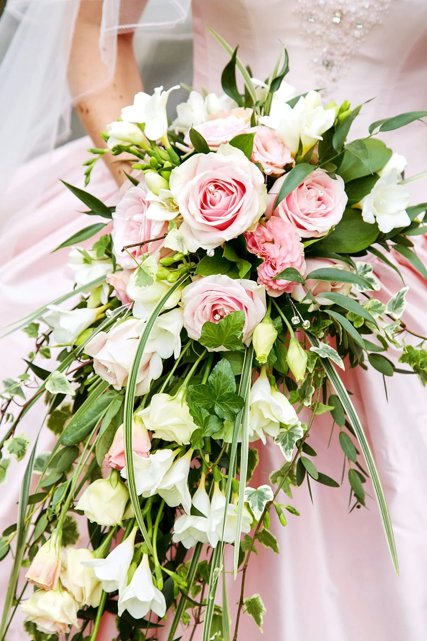 Close-up of a bridal bouquet with pink roses, white flowers, and green foliage, held by a person wearing a pink dress with jewelry details.