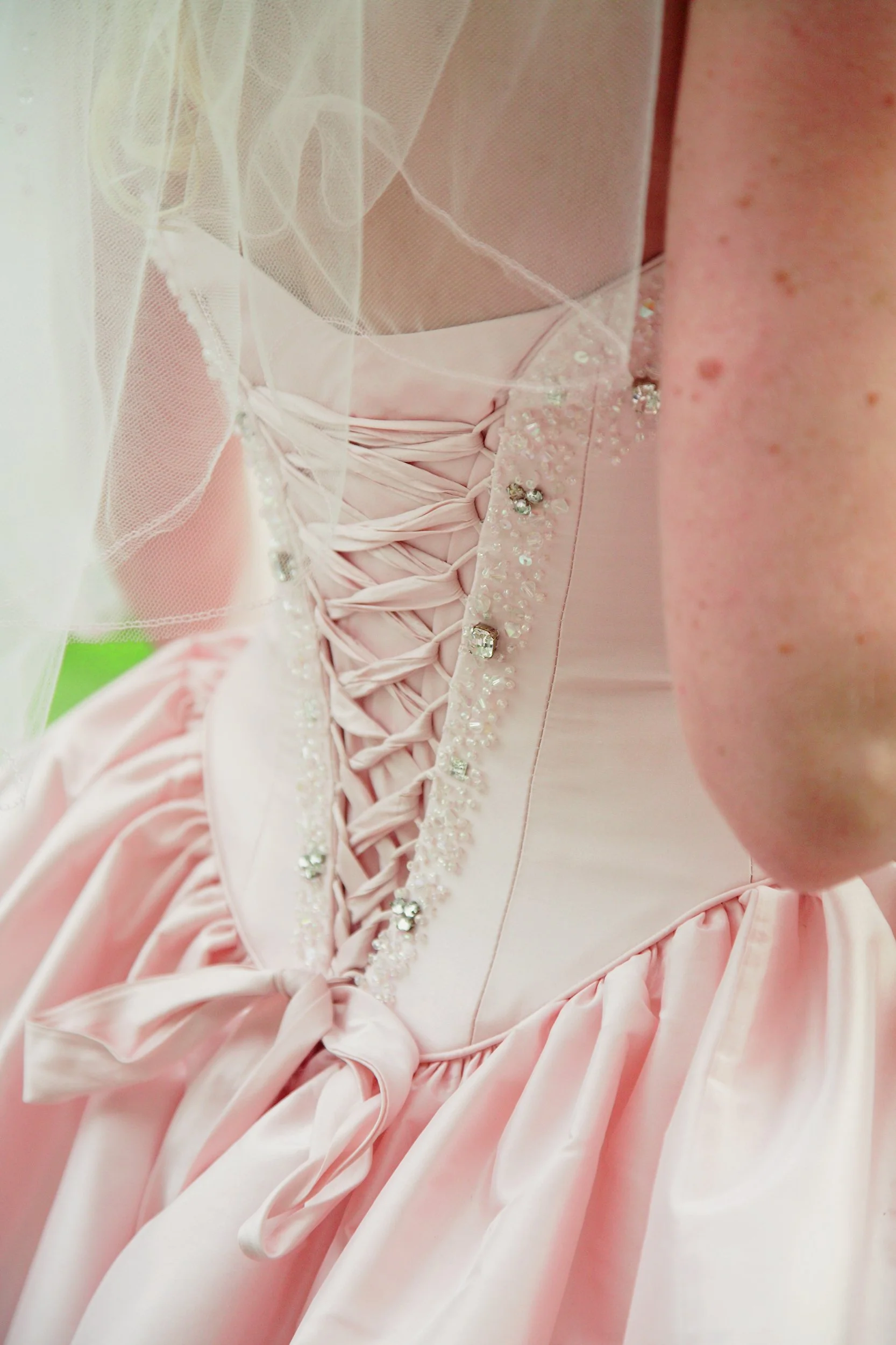 Close-up of a pink dress with satin fabric, lace-up corset back, and rhinestone embellishments, worn by a person with freckles.
