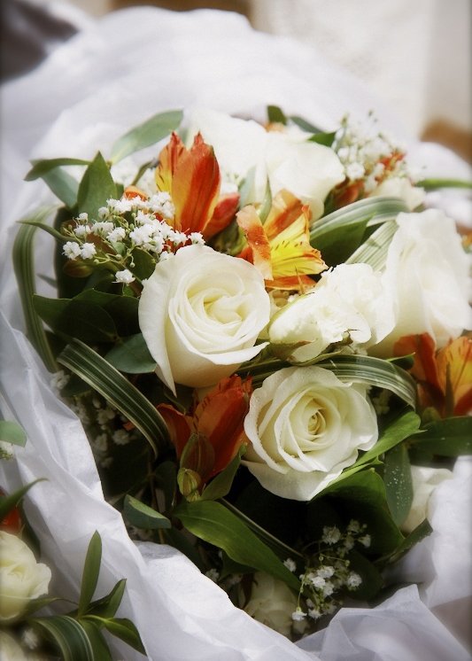 A bouquet of white roses, orange lilies, small white filler flowers, and green leaves wrapped in white paper.