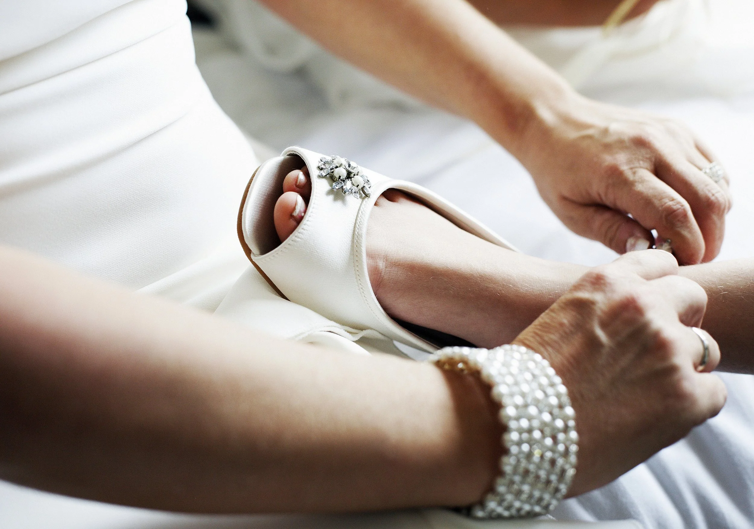 Close-up of a woman's wrist adorned with a pearl and rhinestone bracelet, on a white satin sleeve of a wedding dress, with a man’s hand gently holding her hand.