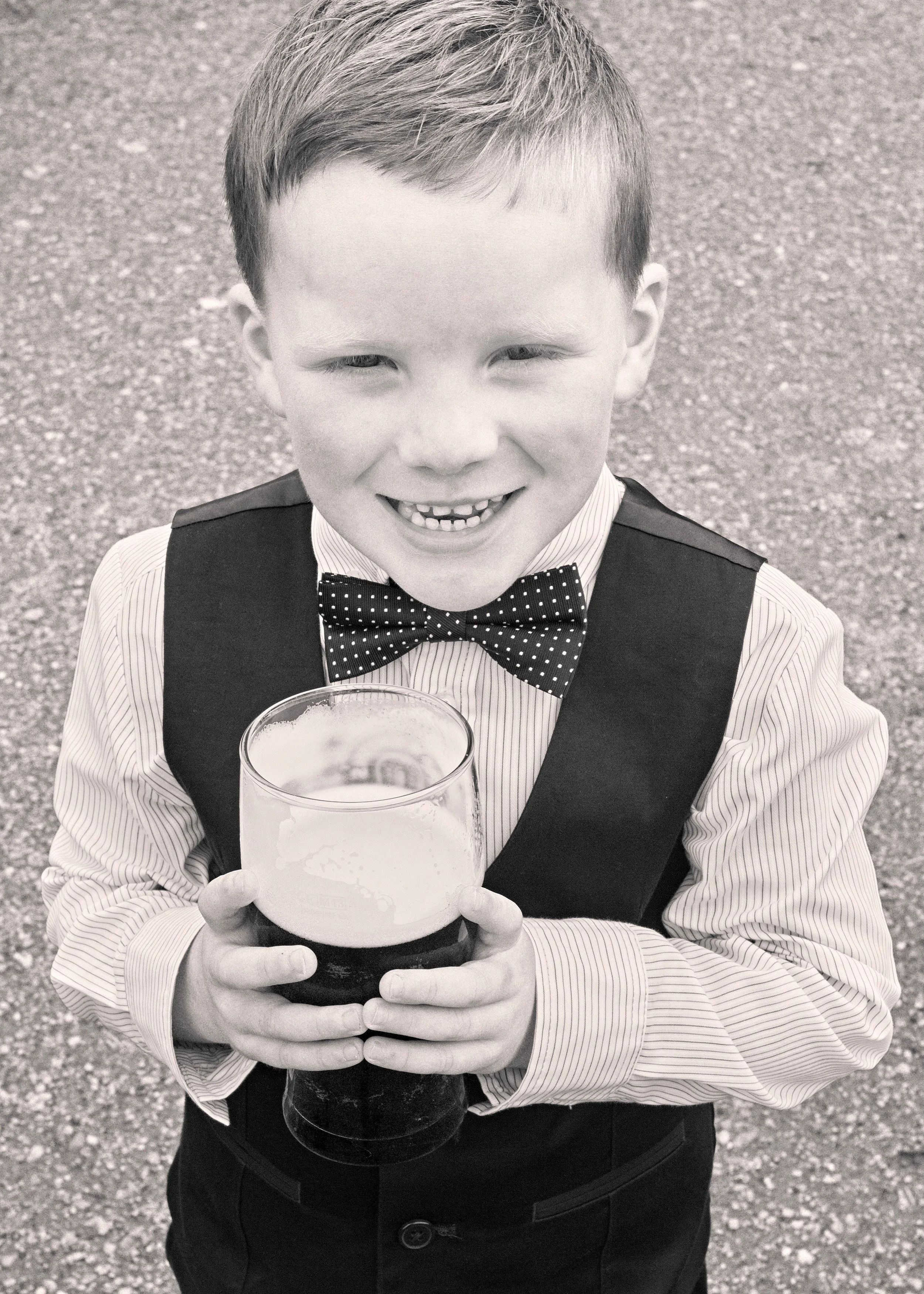 A young boy dressed in formal attire with a bow tie, holding a glass of beer and smiling outside on a gravel surface.