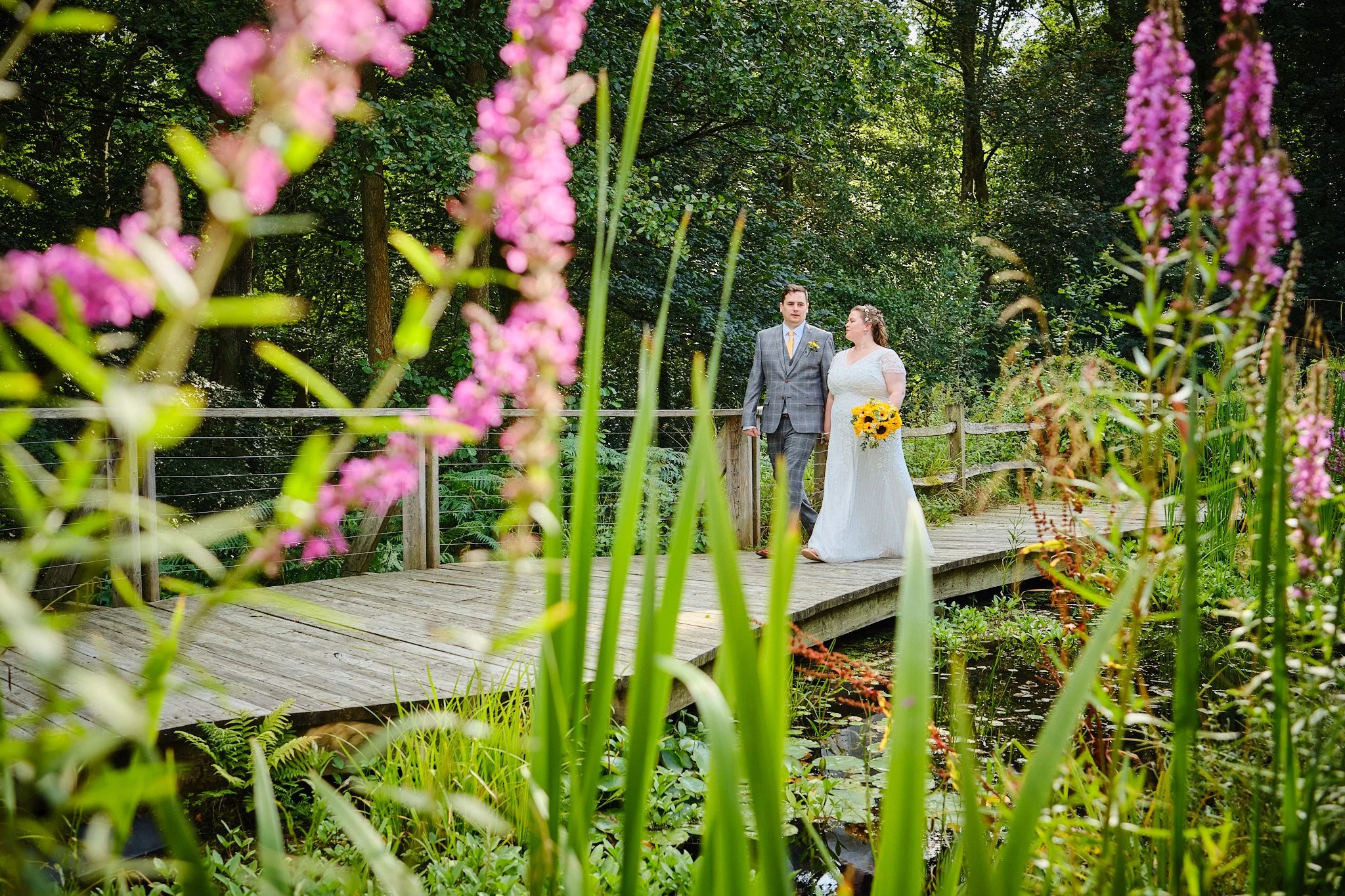 A bride and groom standing on a wooden bridge in a lush, green park surrounded by pink and purple flowers.