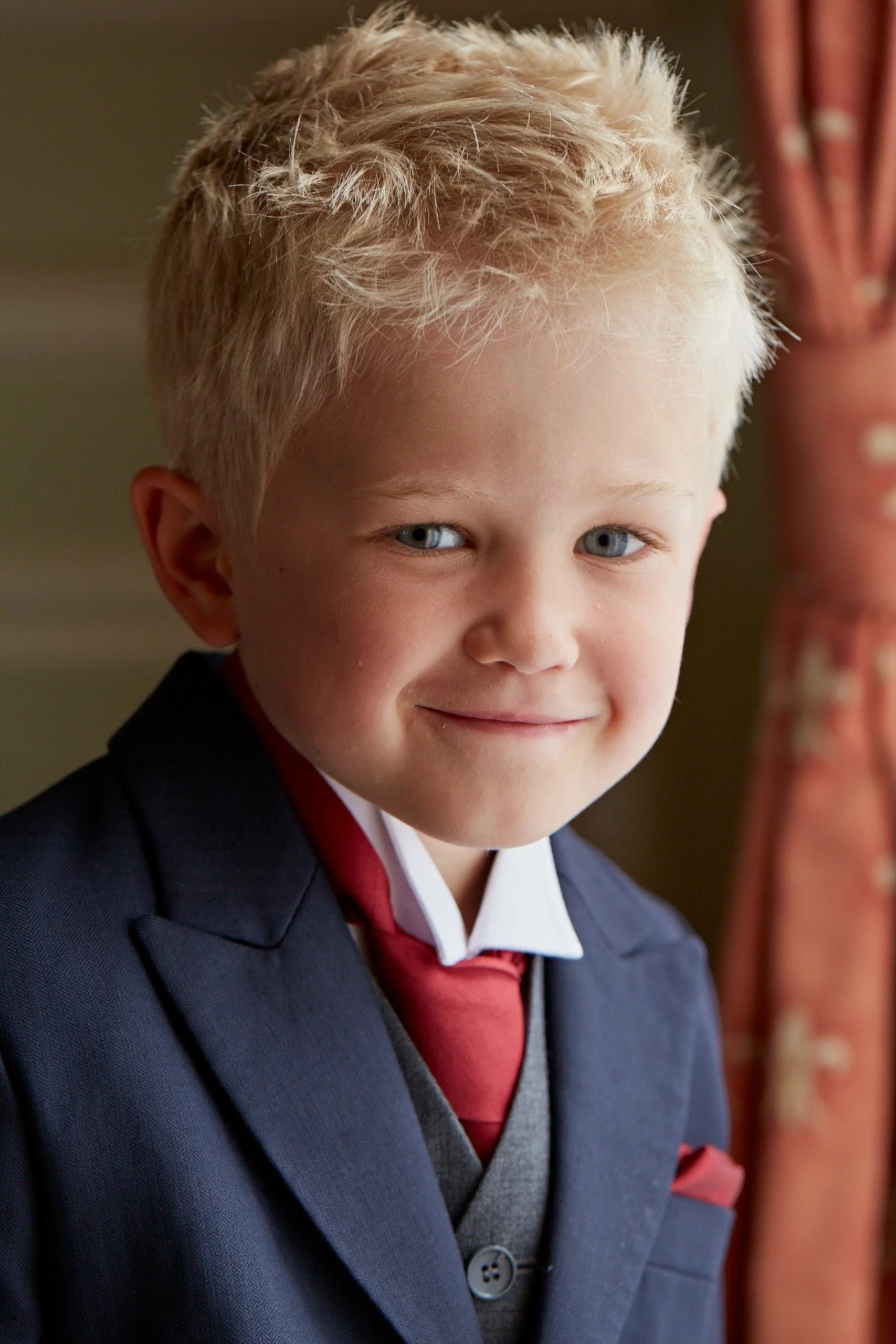 Close-up of a young boy with blonde curly hair, blue eyes, wearing a navy blazer, white shirt, grey sweater vest, and red tie, smiling softly. Background includes a red curtain.