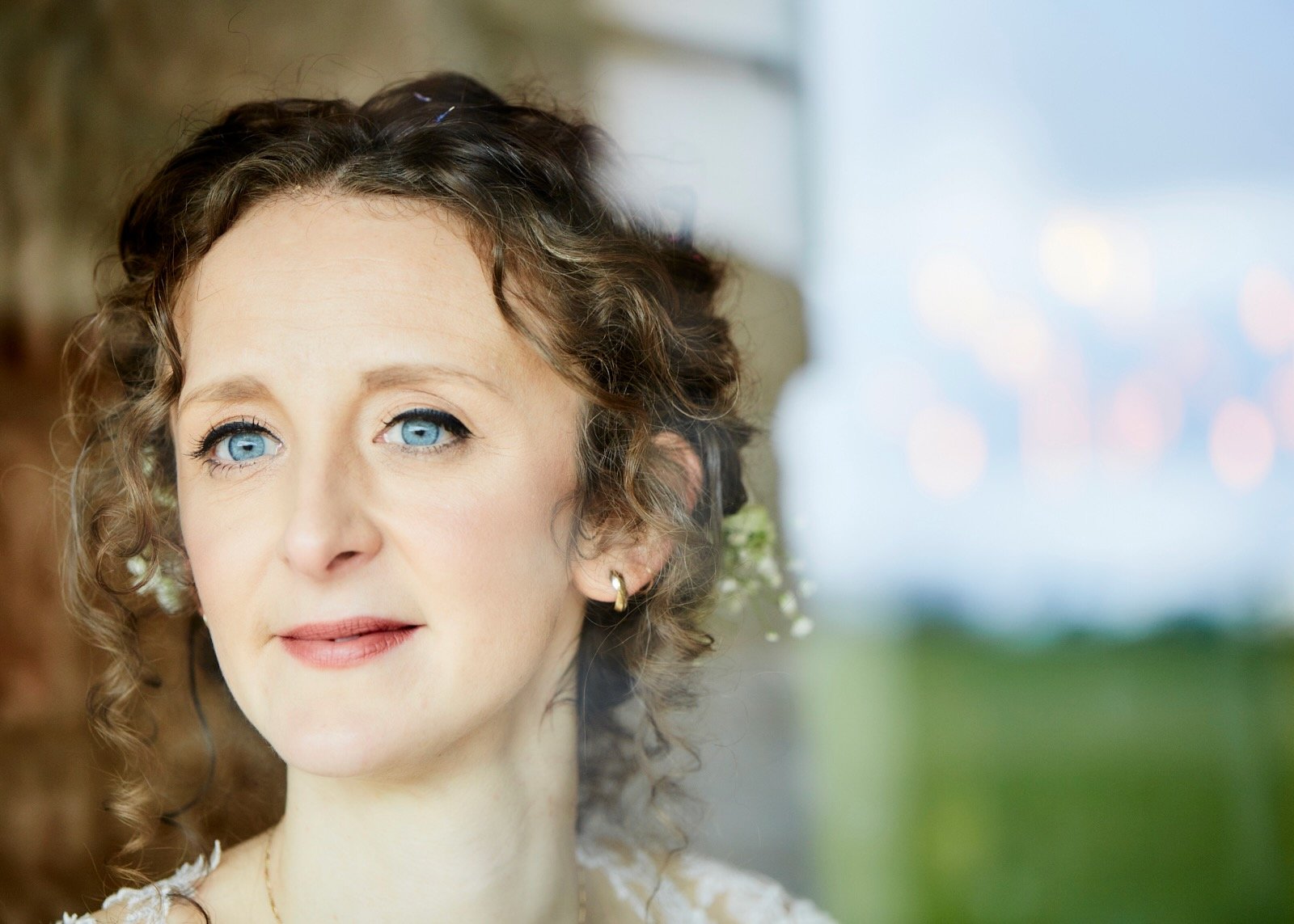 A woman with curly brown hair, blue eyes, and light skin, wearing earrings, standing outdoors with a blurred natural background.