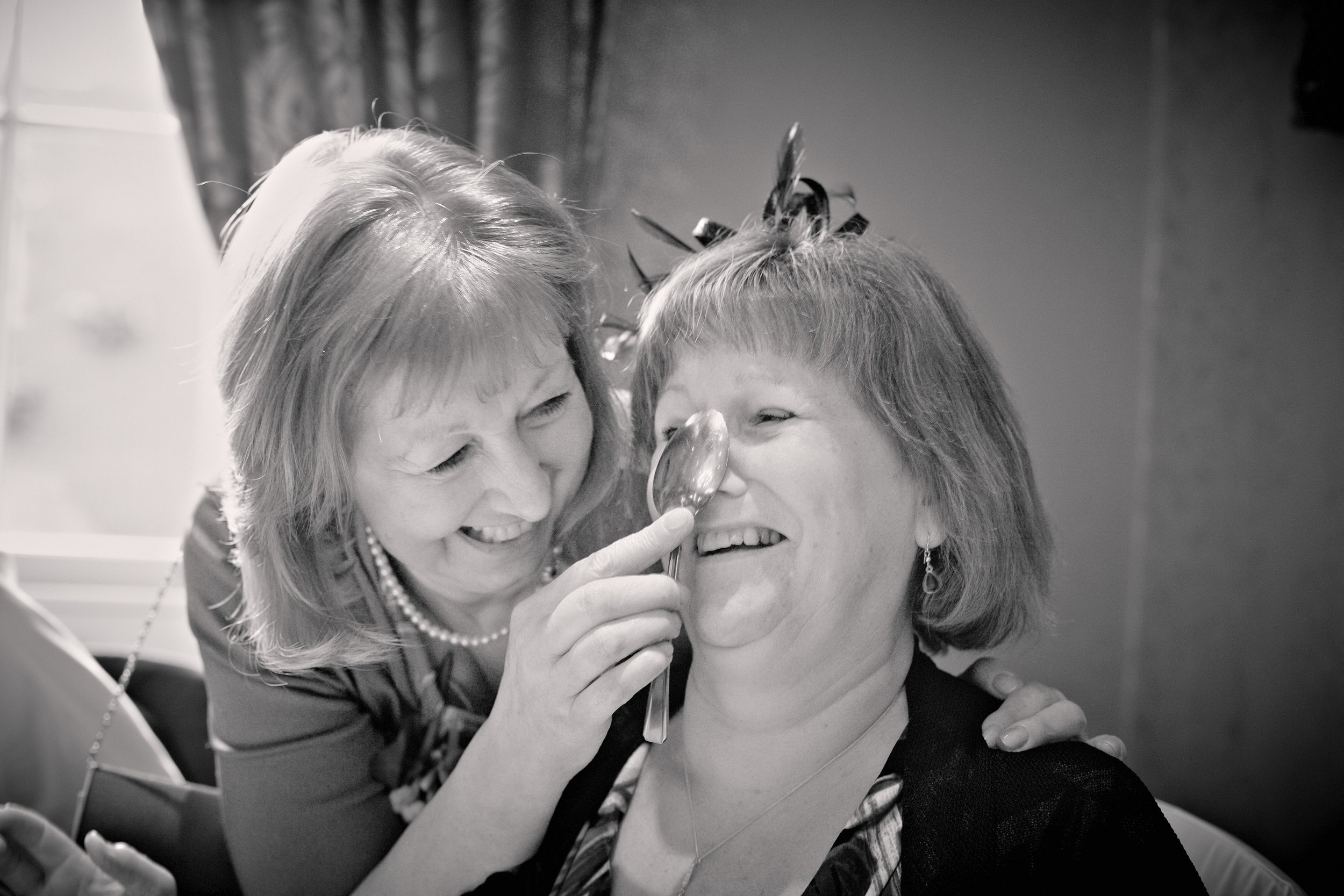 A woman with light-colored hair, wearing jewelry, is smiling and fondling the face of another woman with short hair, who is holding a spoon near her face. They appear to be sharing a joyful moment together.