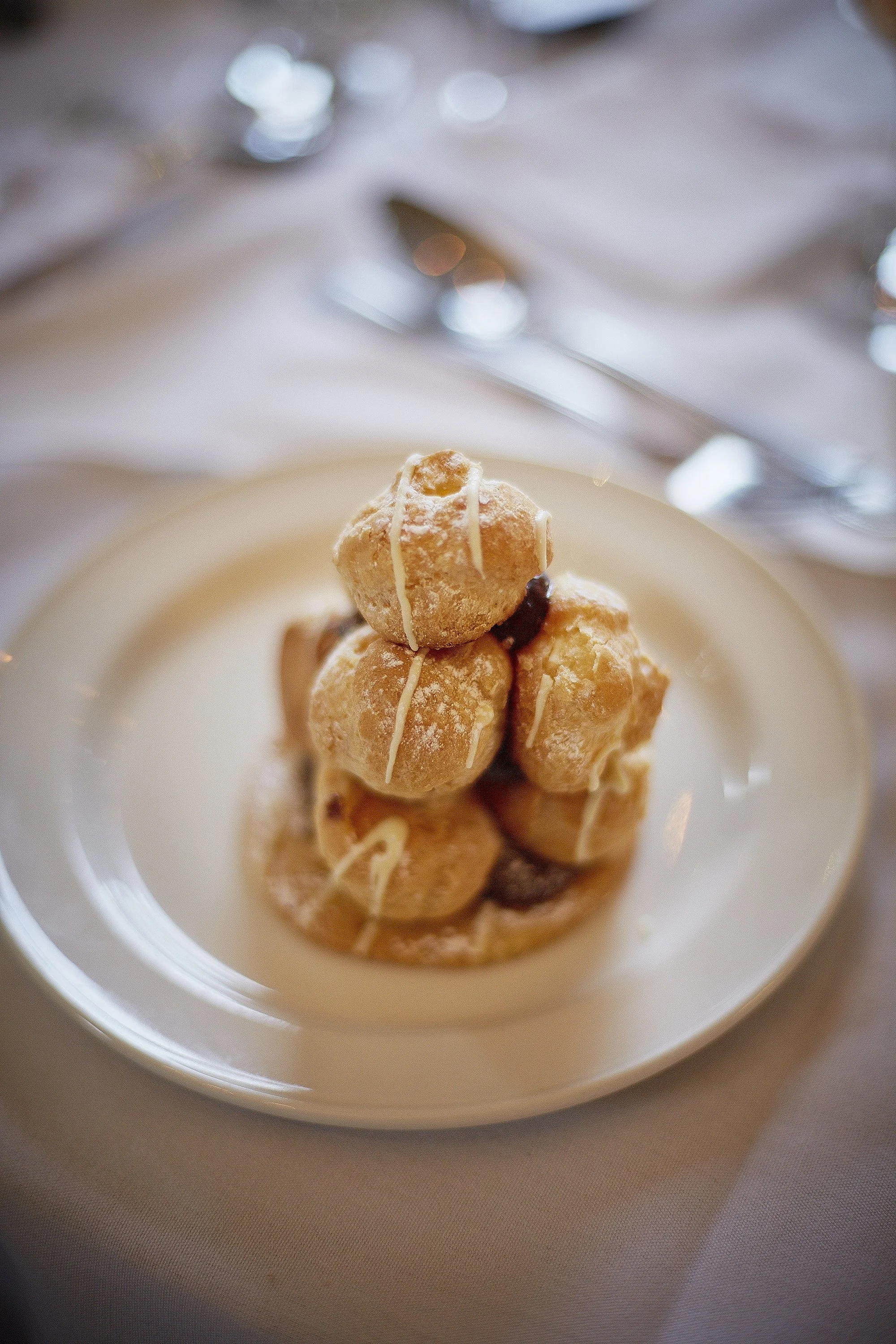 Close-up of a plate of profiteroles drizzled with white chocolate, placed on a table with silverware in the background.
