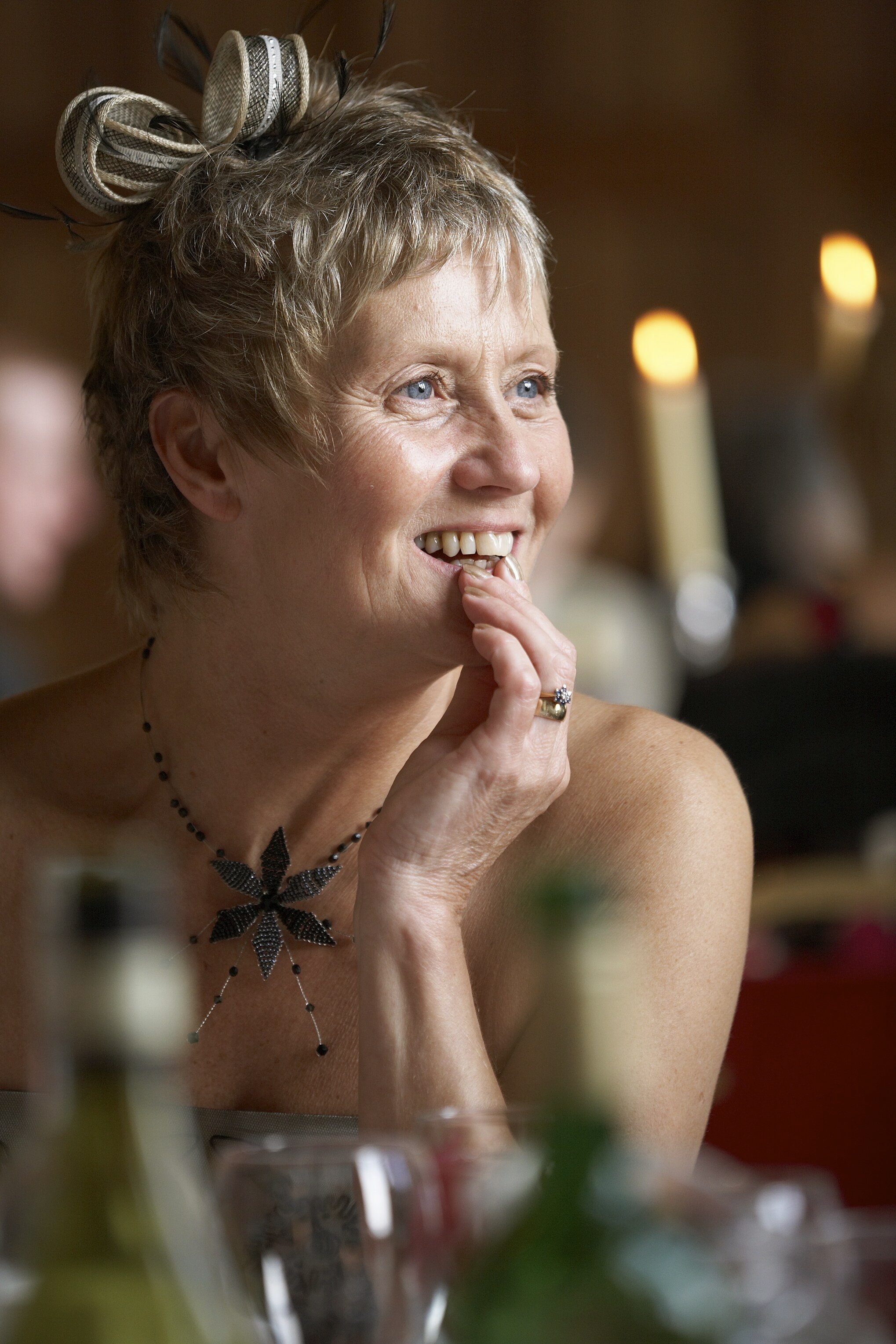 Smiling woman with short blonde hair wearing a black necklace and a large black flower pendant, resting her chin on her hand in a warmly lit indoor setting.