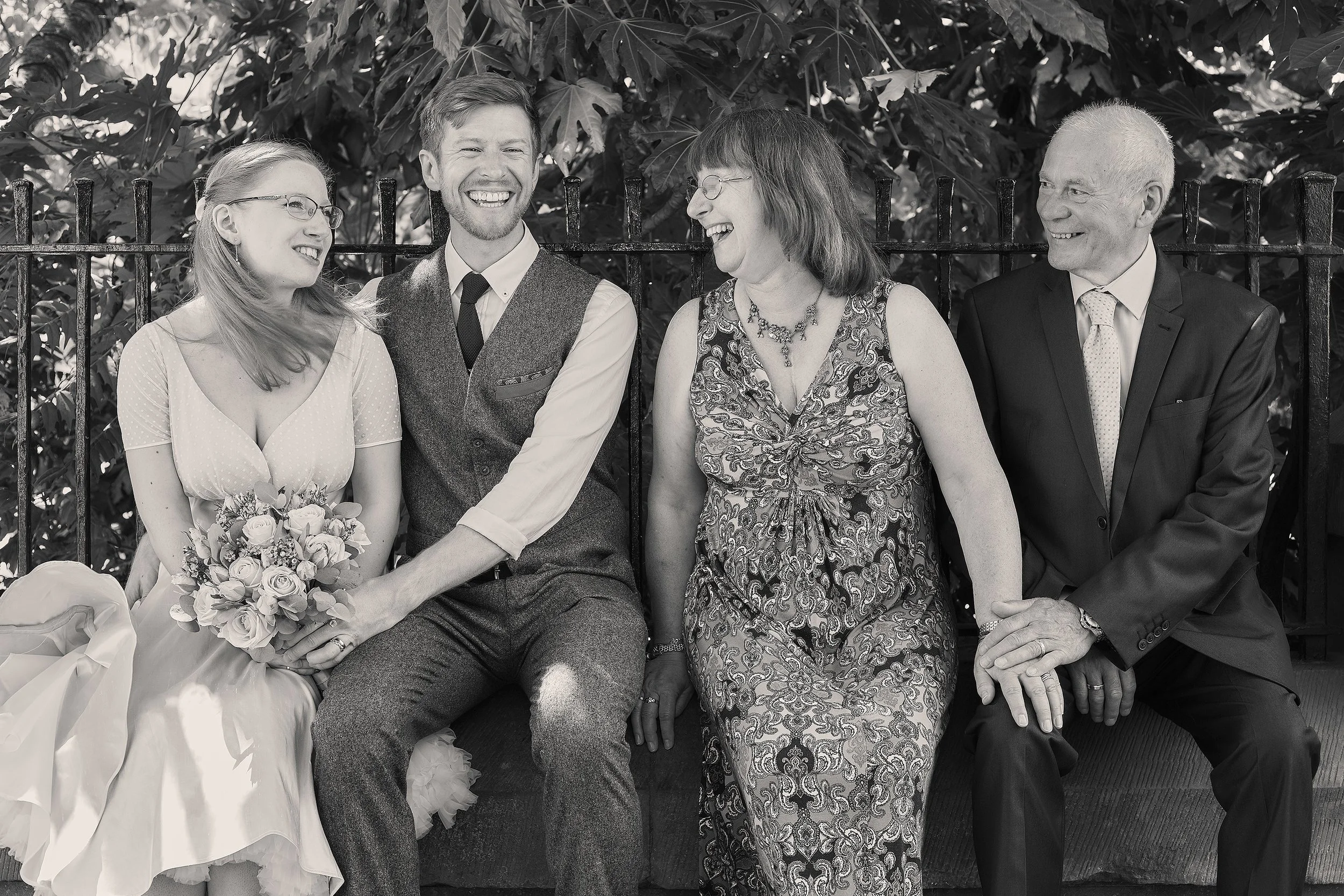 Four people sitting on a bench, happily laughing and smiling, with lush leaves in the background. One woman is holding a bouquet of roses, and the group appears to be enjoying a joyful moment together.