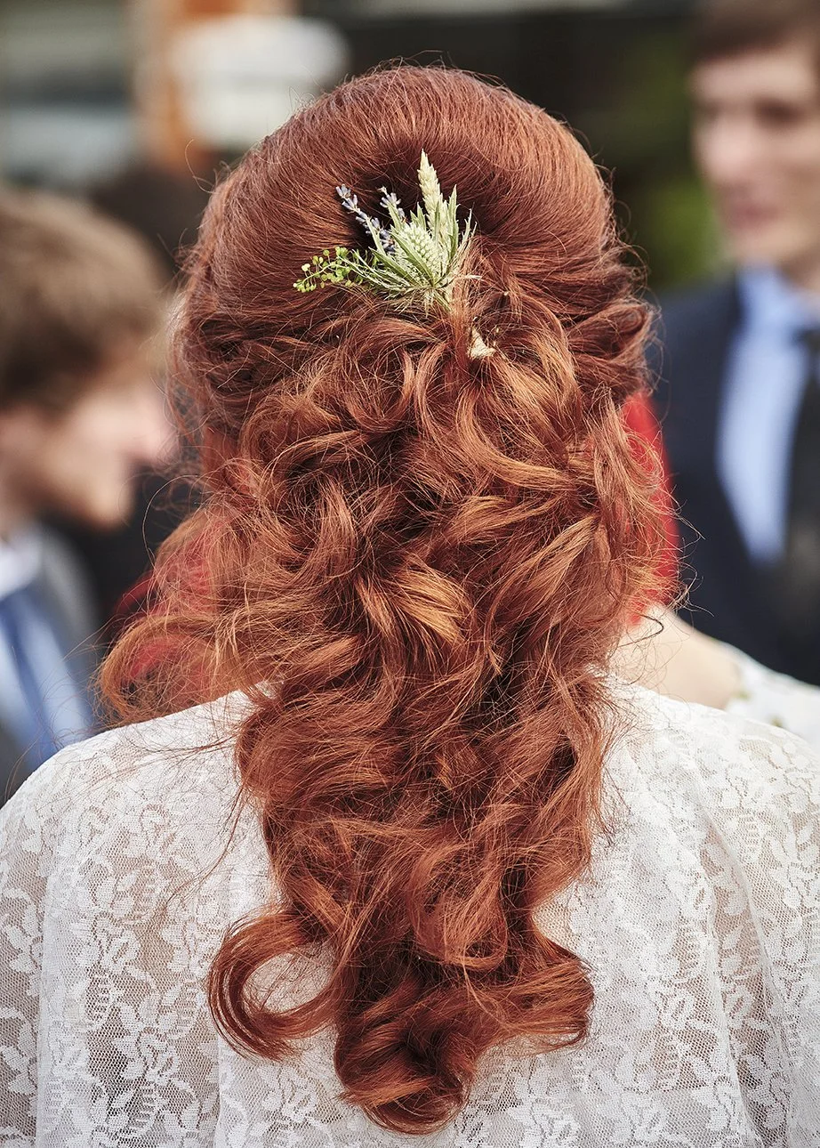 A woman with curly red hair styled in a half-up, half-down hairstyle, adorned with a small floral hair accessory, wearing a white lace garment at an outdoor event.