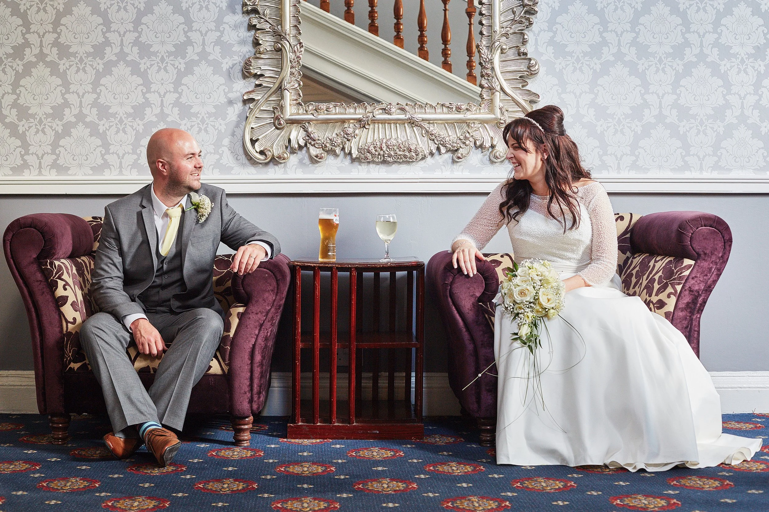 A bride and groom sitting on purple velvet sofas in a decorated room with ornate mirror, wallpaper, and patterned carpet, smiling at each other, with drinks on a small table between them.