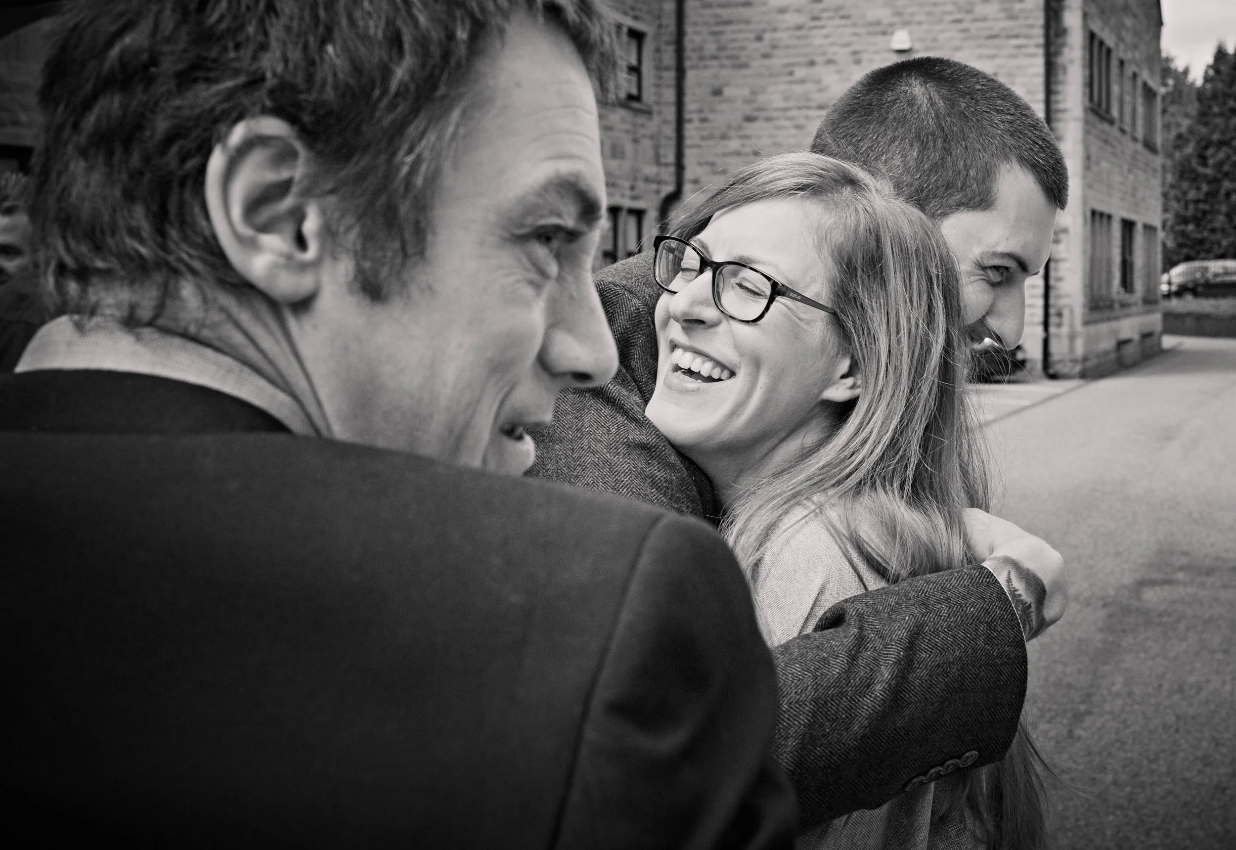 Four people hugging and smiling outdoors, with buildings and trees in the background.