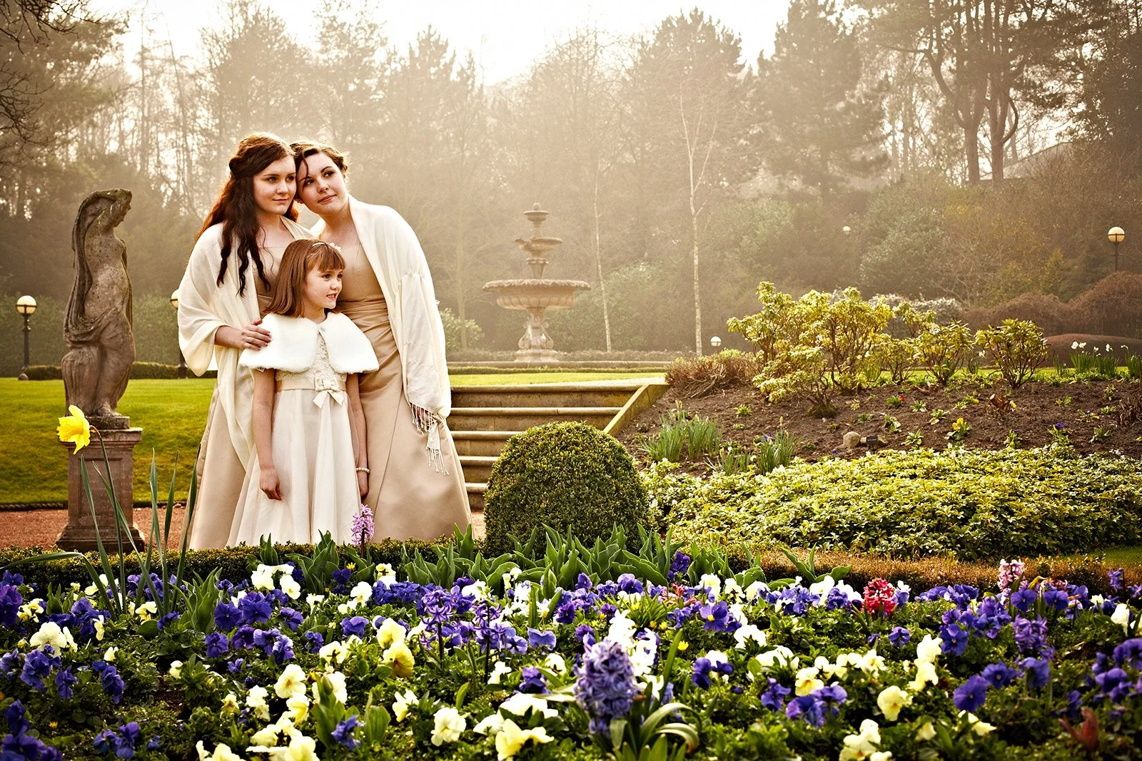 Three women and a girl dressed in white and beige, standing together in a garden with blooming flowers, statues, and a fountain in the background, during a foggy morning or late afternoon.