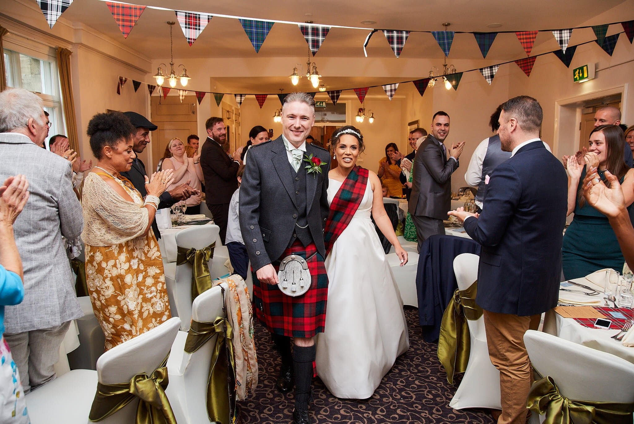 A wedding reception with guests clapping and celebrating. The bride and groom are in the center; the bride wears a white dress with a tartan sash, and the groom wears a gray suit with a tartan boutonniere. The room is decorated with tartan banners an
