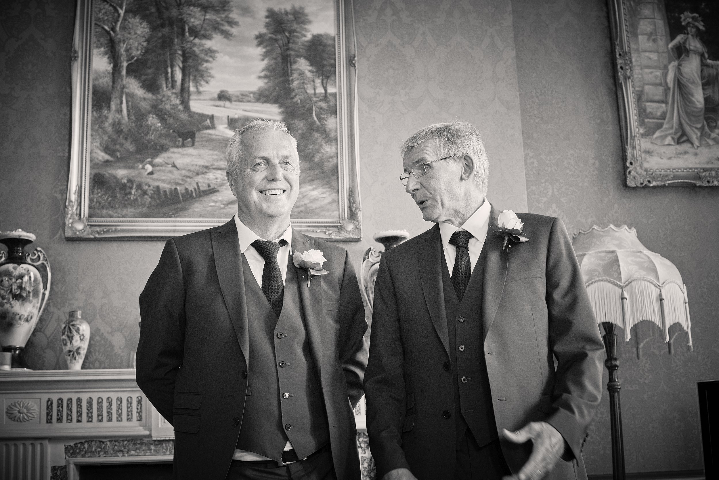 Two elderly men in suits with boutonnières on their lapels are standing in a decorated room, smiling and conversing. The room has ornate wallpaper, framed paintings on the wall, and vases with floral patterns.