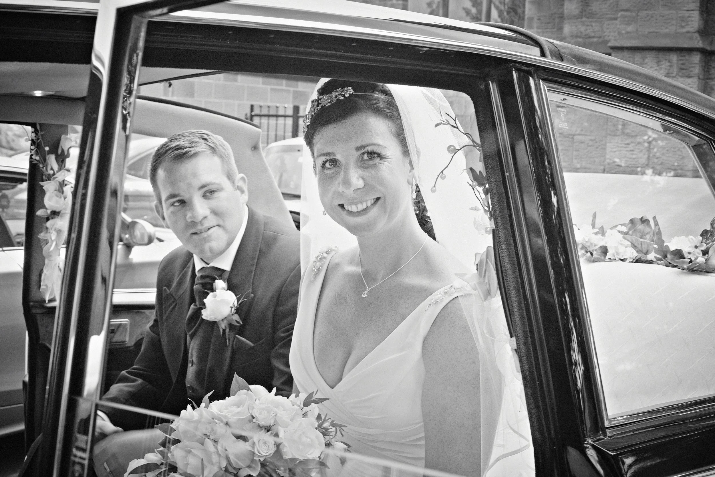 A bride and groom sitting in a vintage car decorated with flowers, with the bride smiling and wearing a veil and tiara, and the groom looking at her.