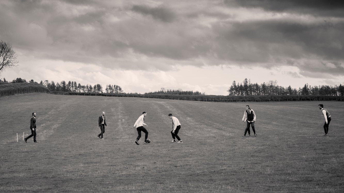 Six men in suits and masks playing soccer on a grassy field under a cloudy sky.
