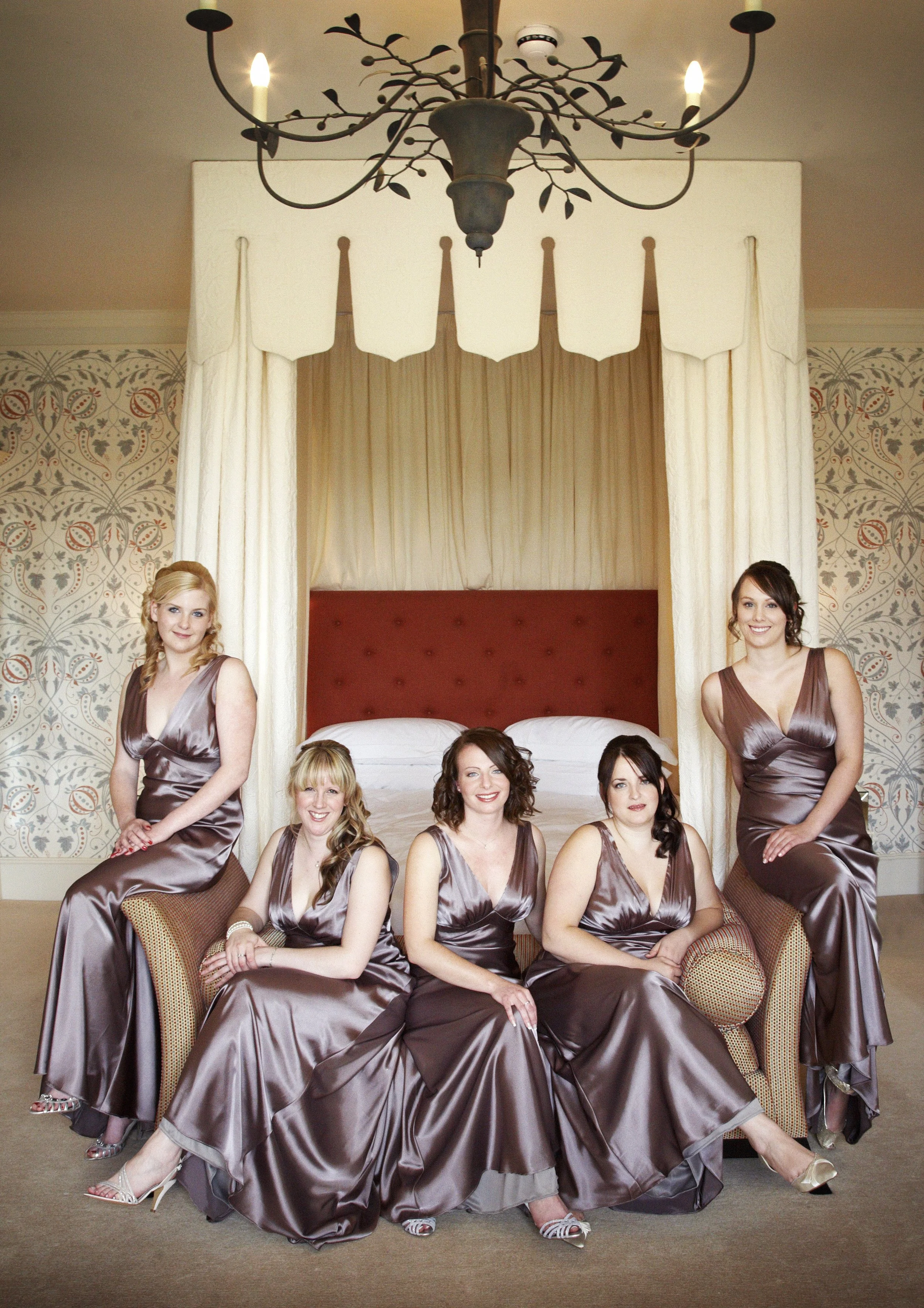 A group of seven women in matching silver satin dresses posing in a hotel room with a bed, chandelier, and wallpaper. Most women are seated on chairs or couches, with two women standing.