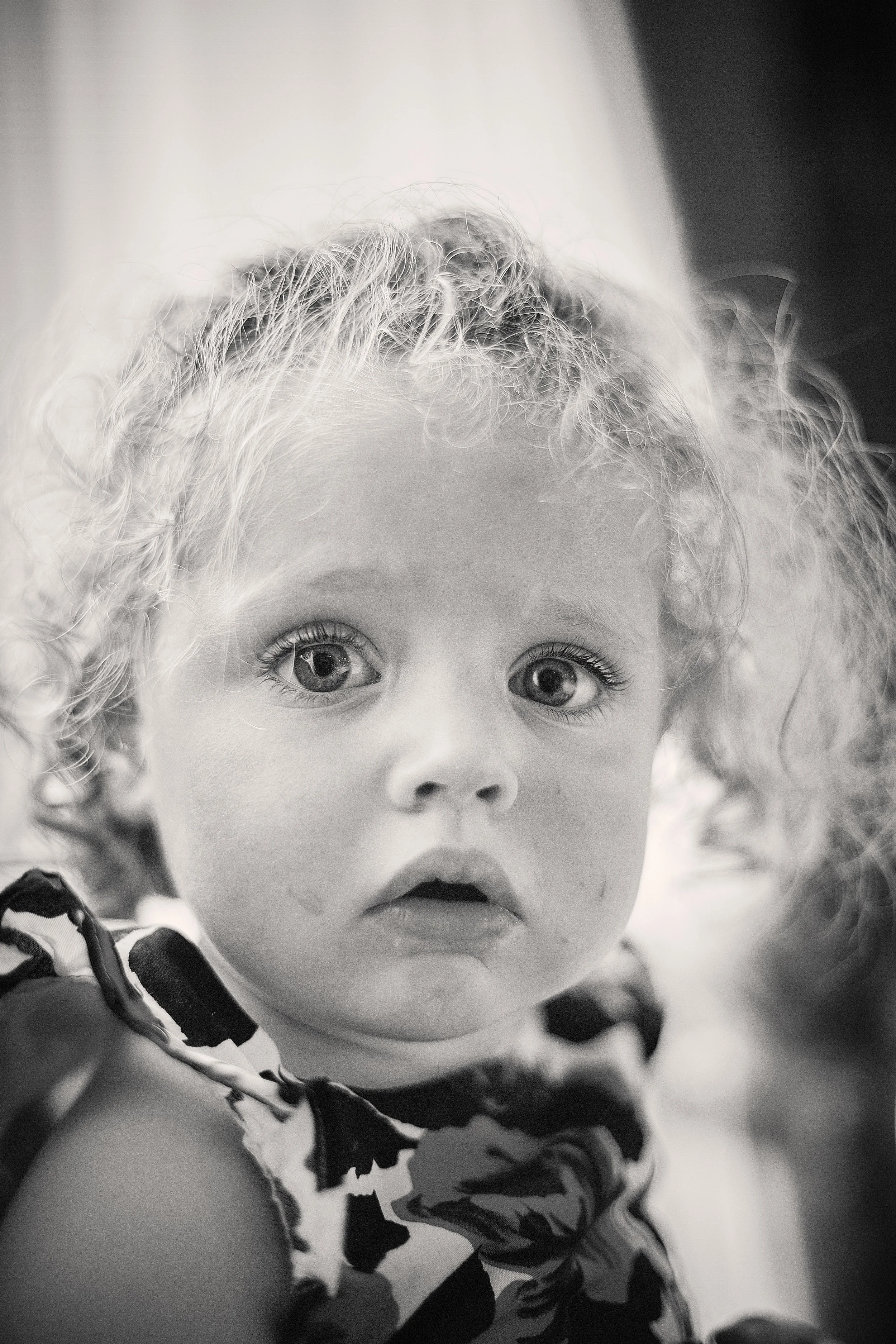 Close-up black and white photo of a young child's face, highlighting large, expressive eyes, curly hair, and wearing a patterned shirt.
