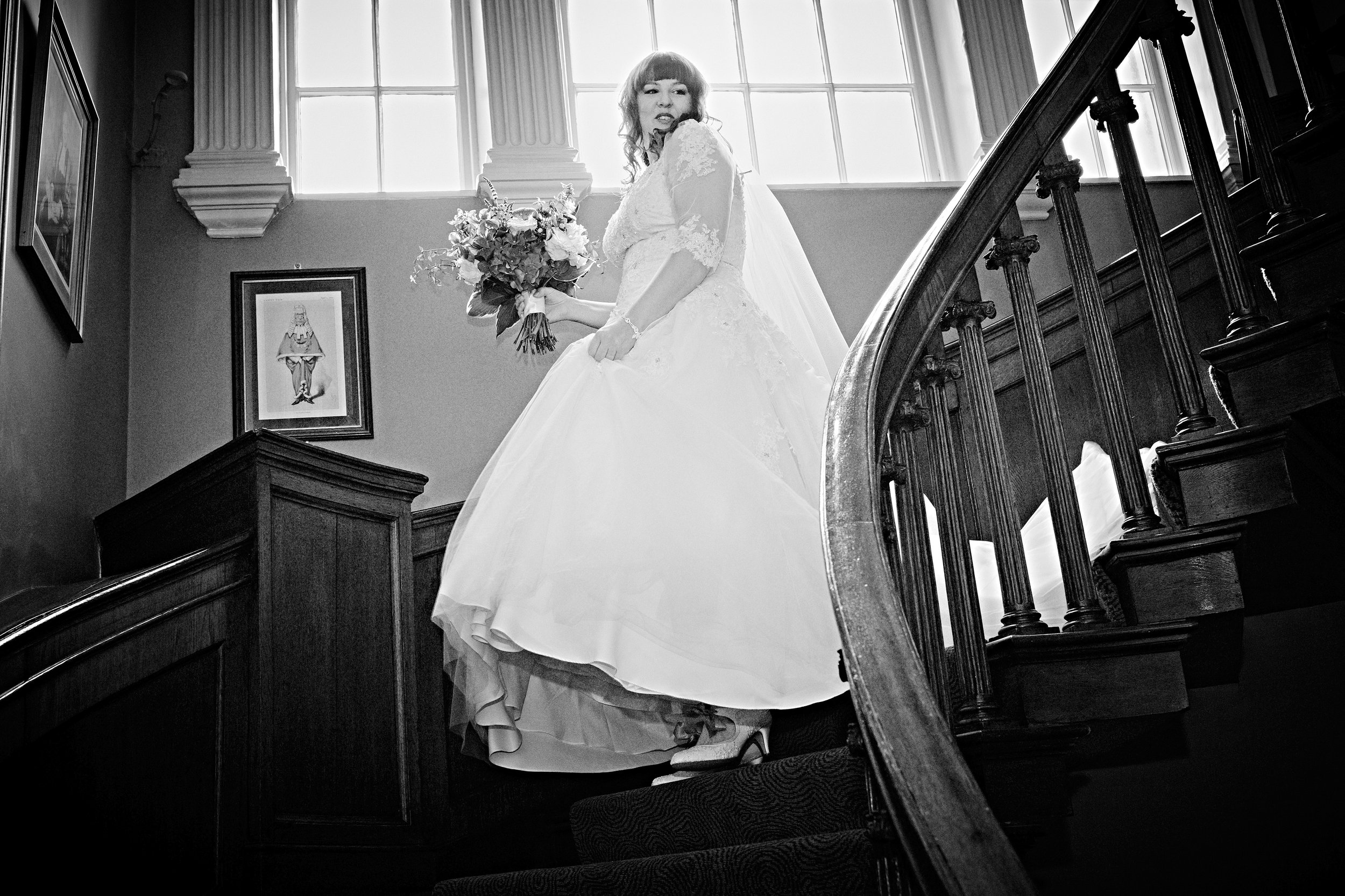 Black and white photo of a bride holding a bouquet of flowers on a staircase inside a building with large windows and framed artwork on the wall.