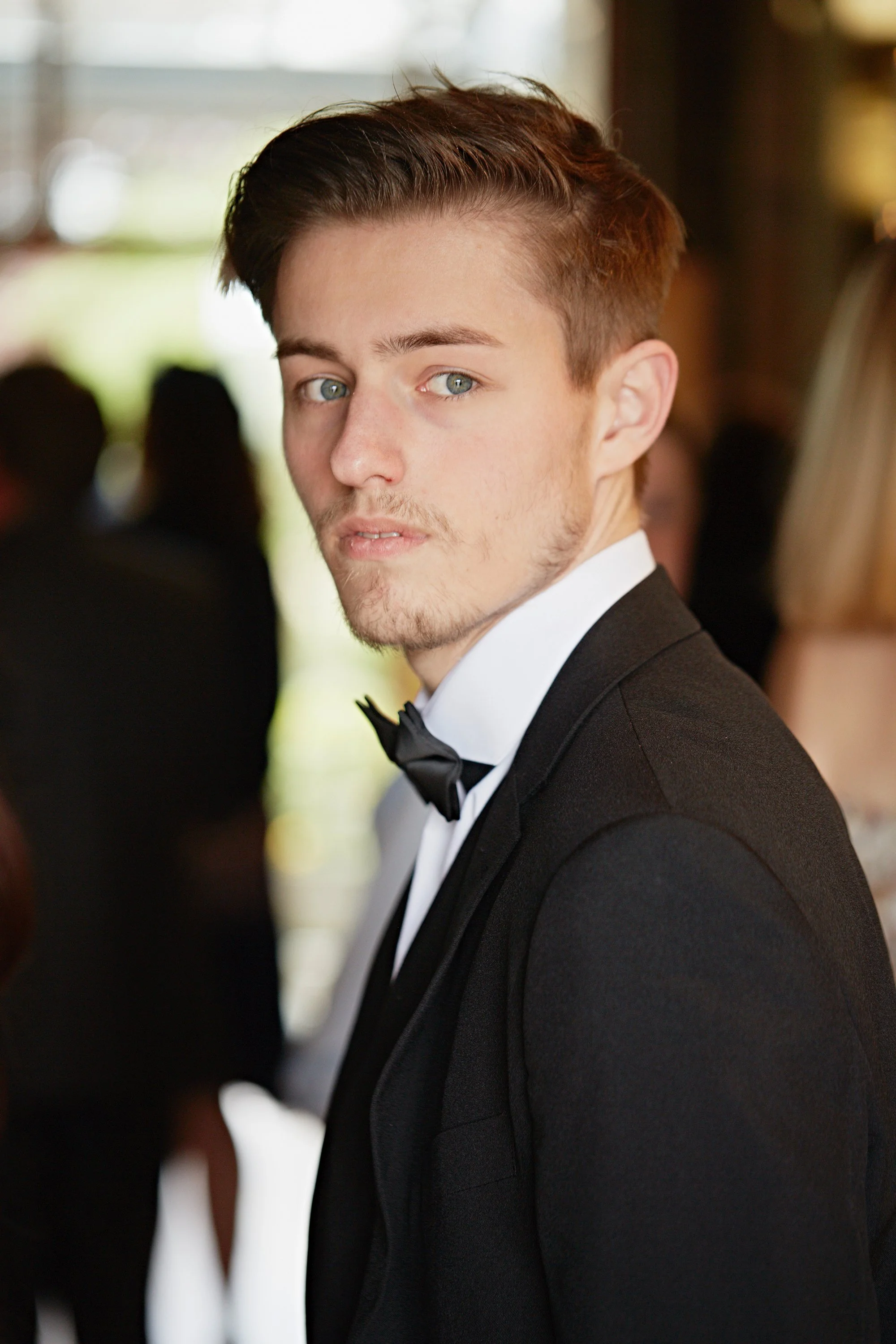 A young man with light skin, short brown hair, and blue eyes is dressed in a black tuxedo with a white dress shirt and black bow tie. He is looking slightly over his shoulder, in a formal setting with blurred people in the background.