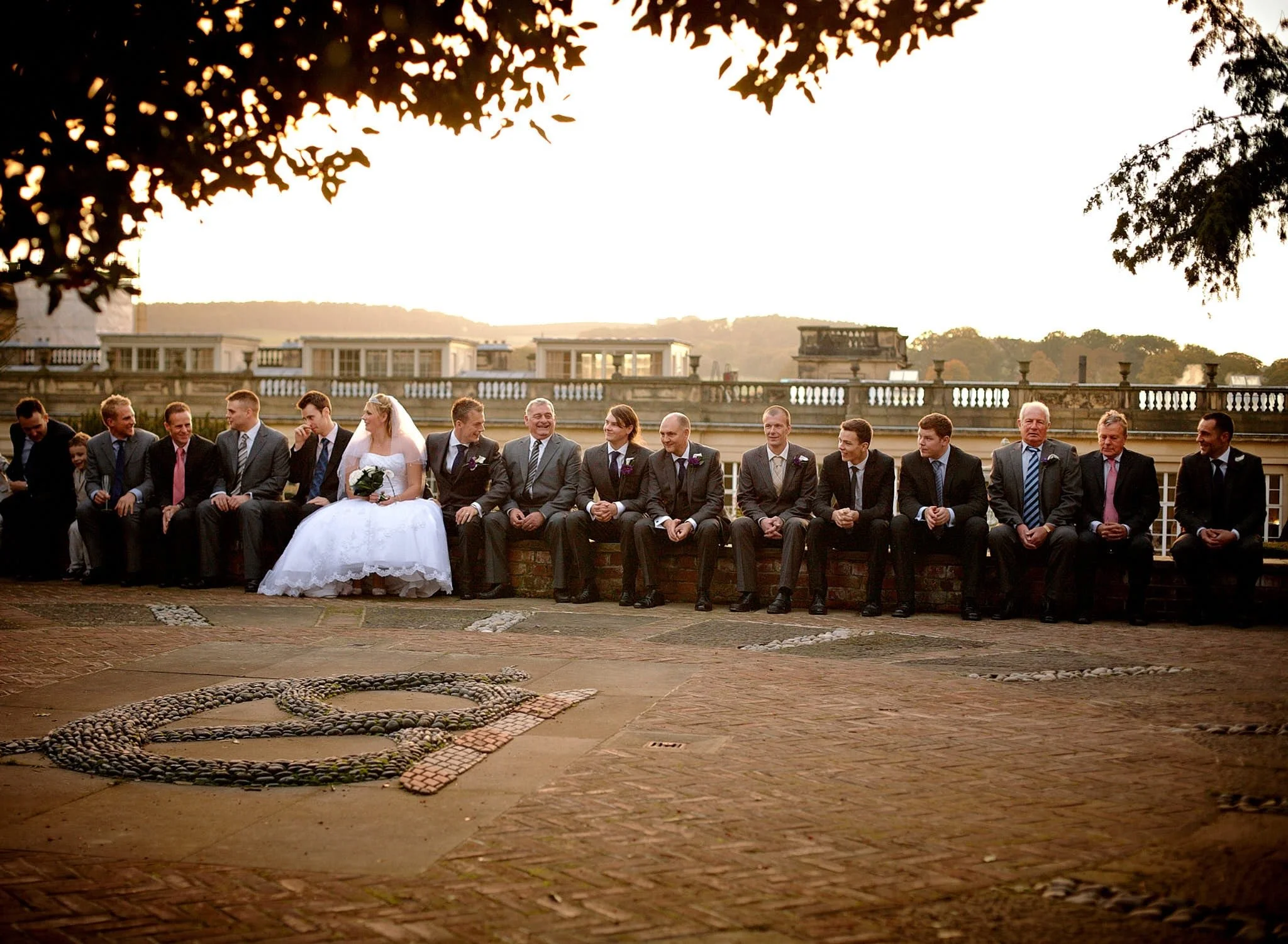 A group of people dressed in formal attire, including a bride in a wedding gown and a groom, sitting outdoors in front of a grand, historic building during sunset.