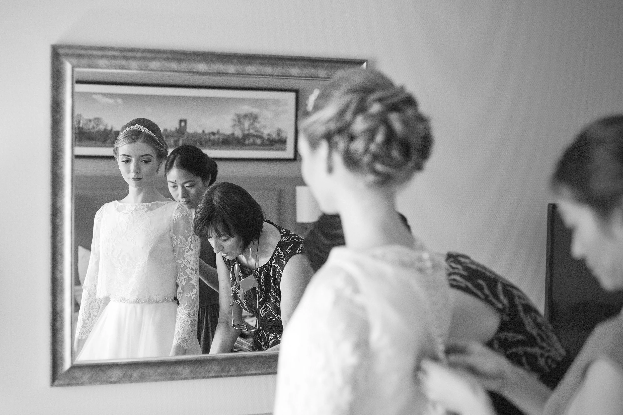 Black and white photograph of a bride in a lace wedding dress looking into a mirror, with two women helping her. One woman has short dark hair and is wearing a patterned dress, the other woman has short hair and glasses, and is wearing a dark dress. 