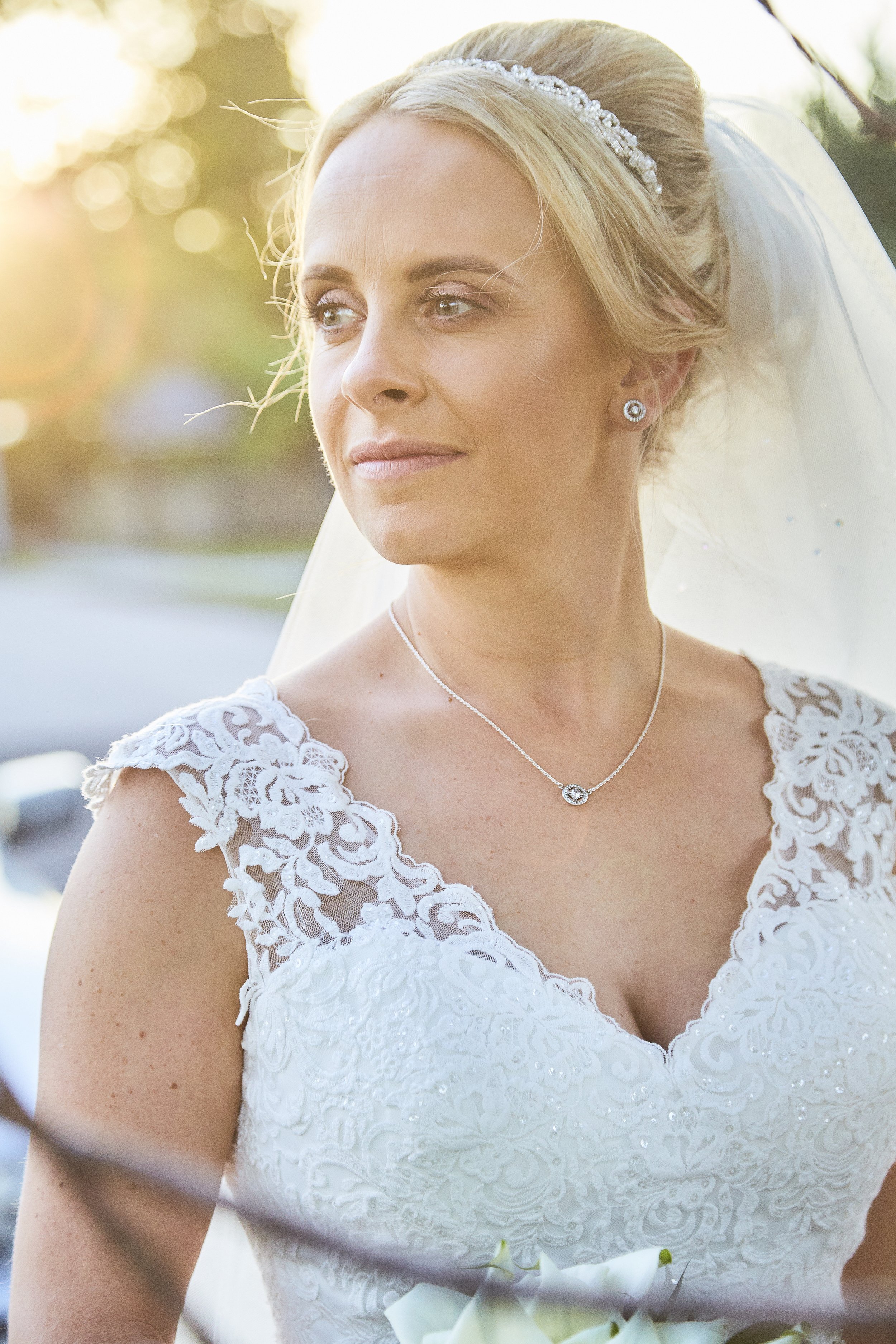 Close-up of a bride with blonde hair styled up, wearing a tiara, pearl earrings, a delicate necklace, and a white lace wedding dress with floral embroidery, standing outdoors during sunset.