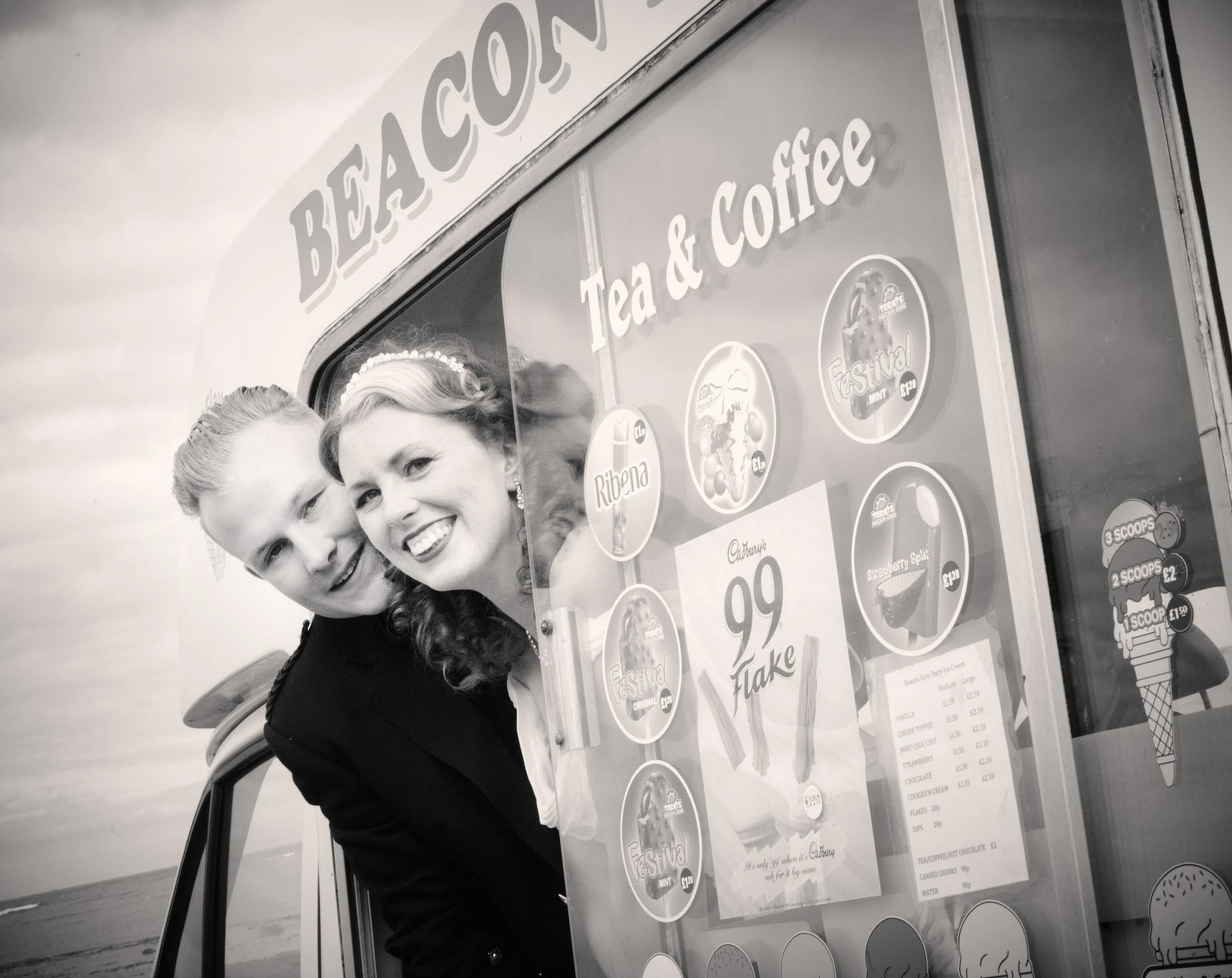 A woman and a young girl smiling at the camera beside a colorful ice cream stand with a menu and promotional posters on the side.