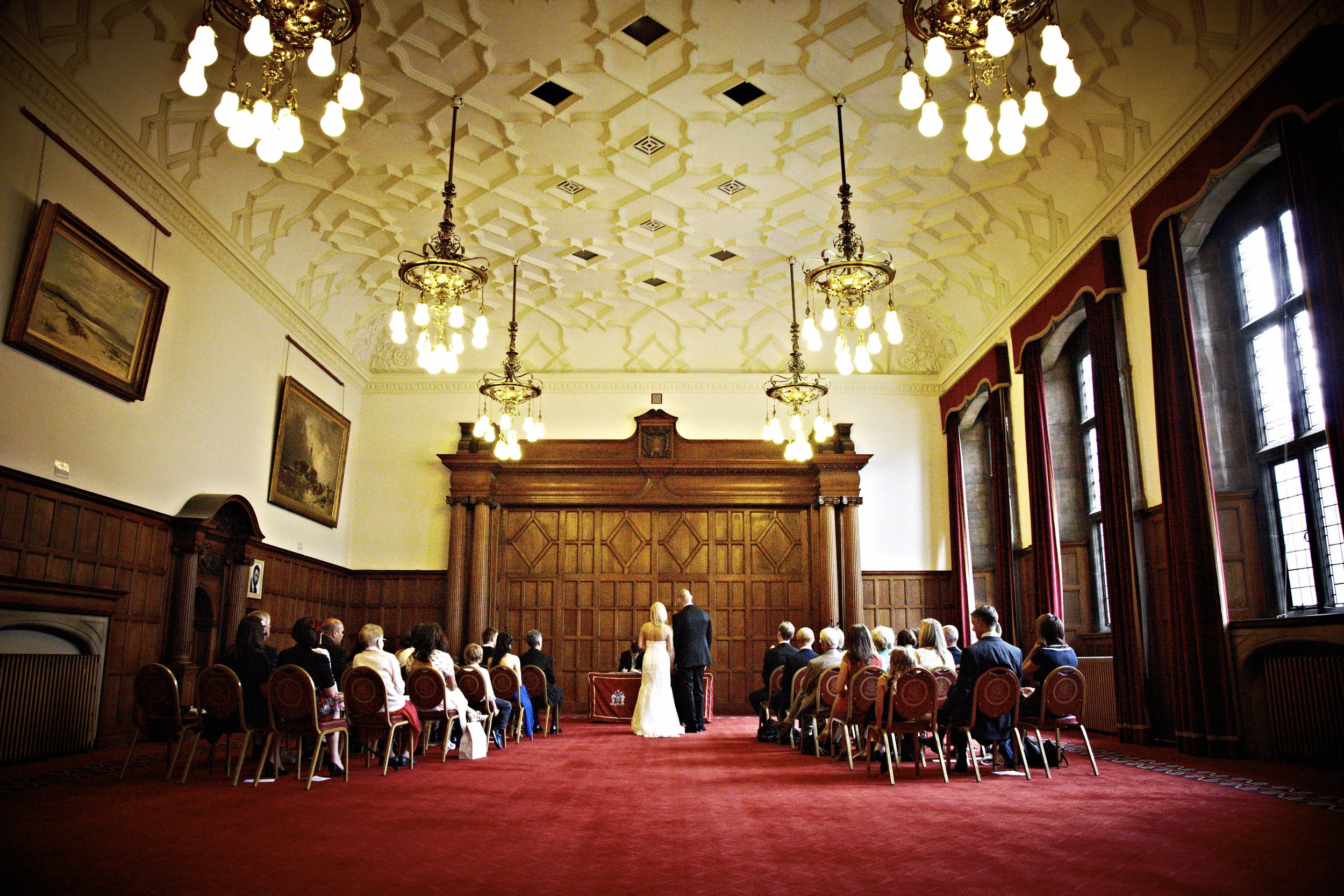 Wedding ceremony in a grand, historic room with high ornate ceiling, chandeliers, and large windows. Guests are seated in rows of chairs facing the altar, where a couple is standing with an officiant. The room has dark wood paneling and framed painti