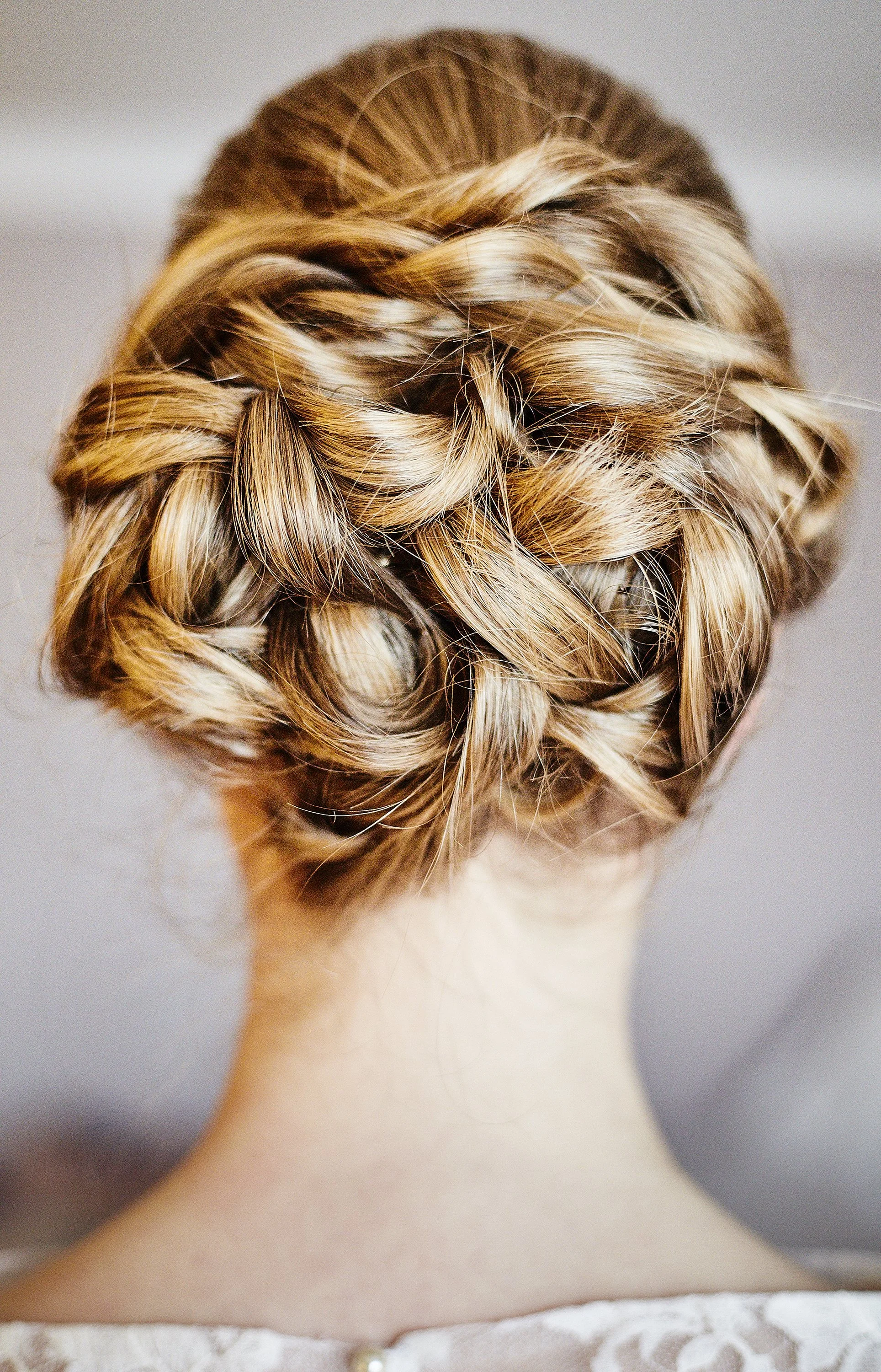 Back view of a woman with intricately styled blonde hair in a braided bun, wearing a white lace top.