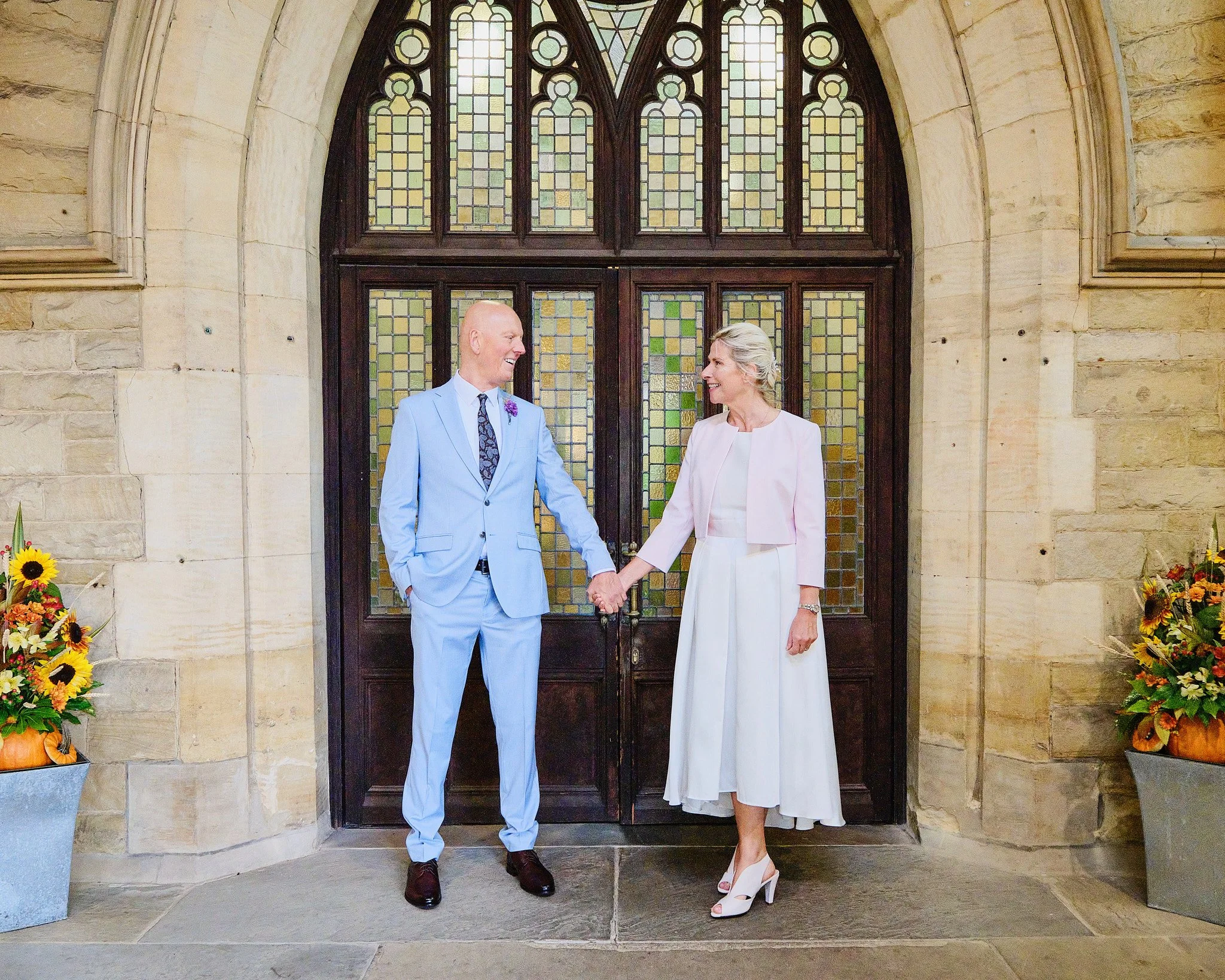 A man and woman holding hands in front of a church door; the man is dressed in a light blue suit and the woman in a white dress, both are smiling and looking at each other, with flower arrangements on either side of the door.