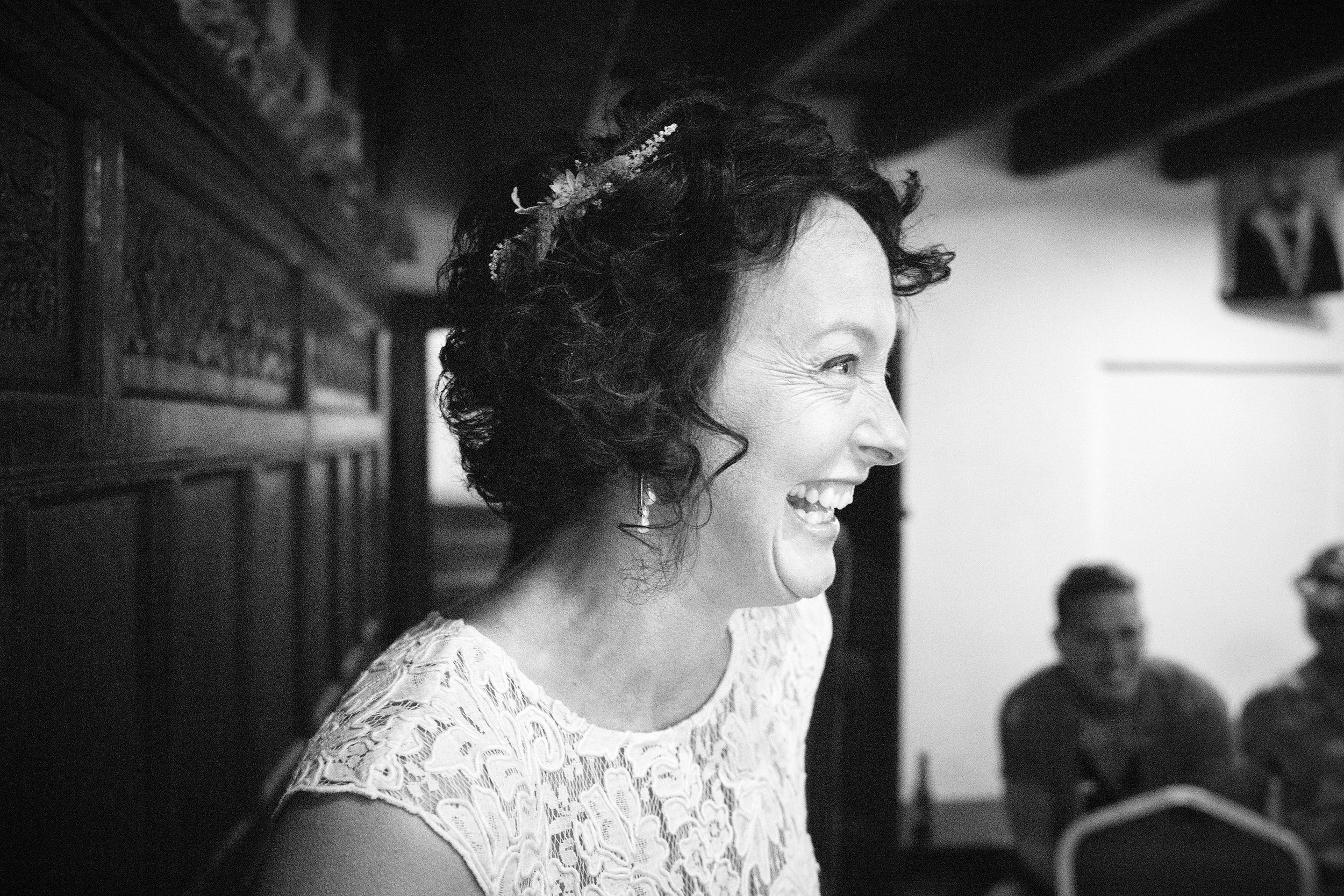 A woman with dark, curly hair adorned with a floral headpiece, smiling and laughing while wearing a lace dress at an indoor event.