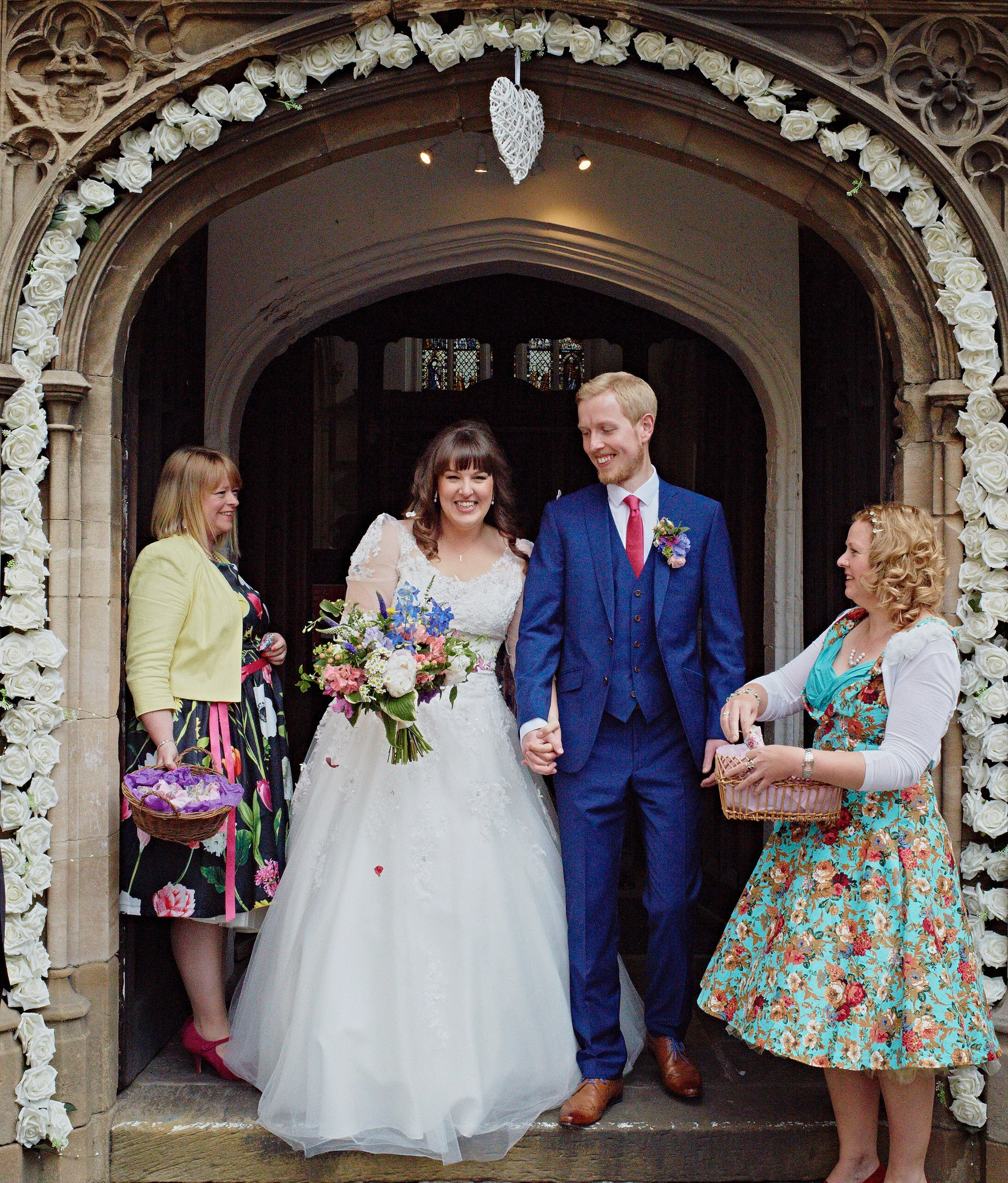 A wedding celebration taking place in front of a church doorway decorated with white roses. The bride in a white gown and the groom in a blue suit are smiling and holding hands. Several guests, mostly women, are dressed in colorful attire, some holdi