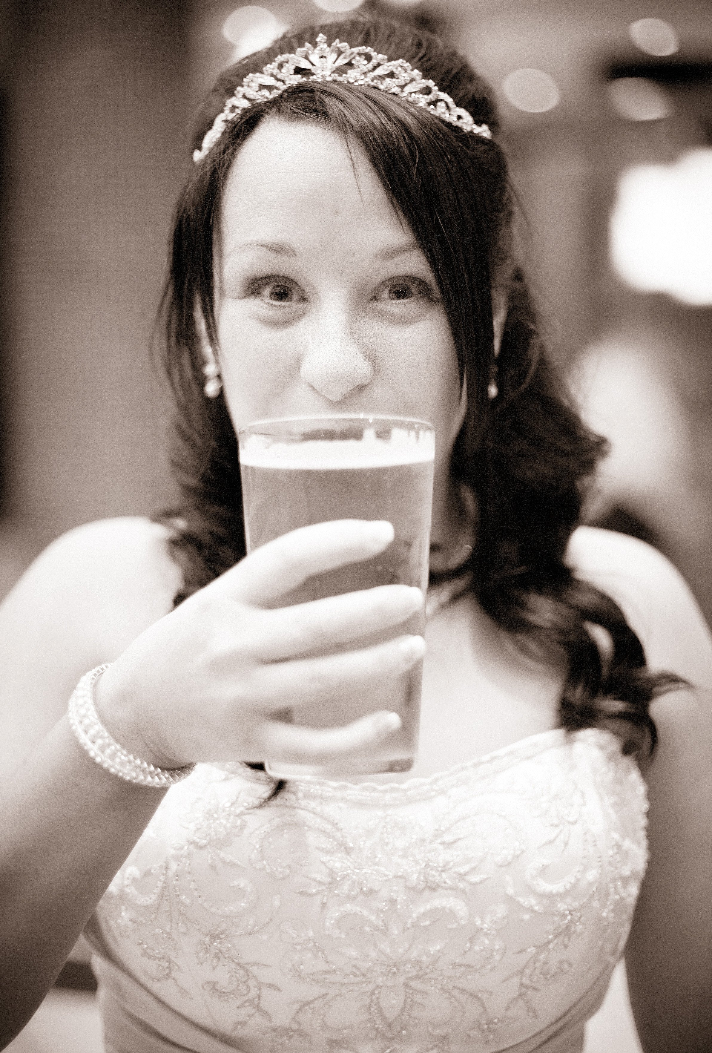 A woman wearing a wedding dress and tiara, holding a glass of wine or champagne, smiling with partial face visible.