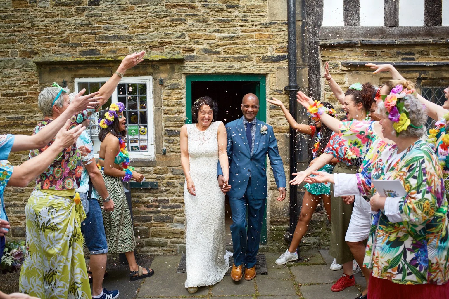 A newlywed couple, holding hands, celebrating outdoors as friends throw confetti around them. The bride is wearing a white lace dress and the groom a blue suit. They are standing in front of a stone building with a window, smiling joyfully.