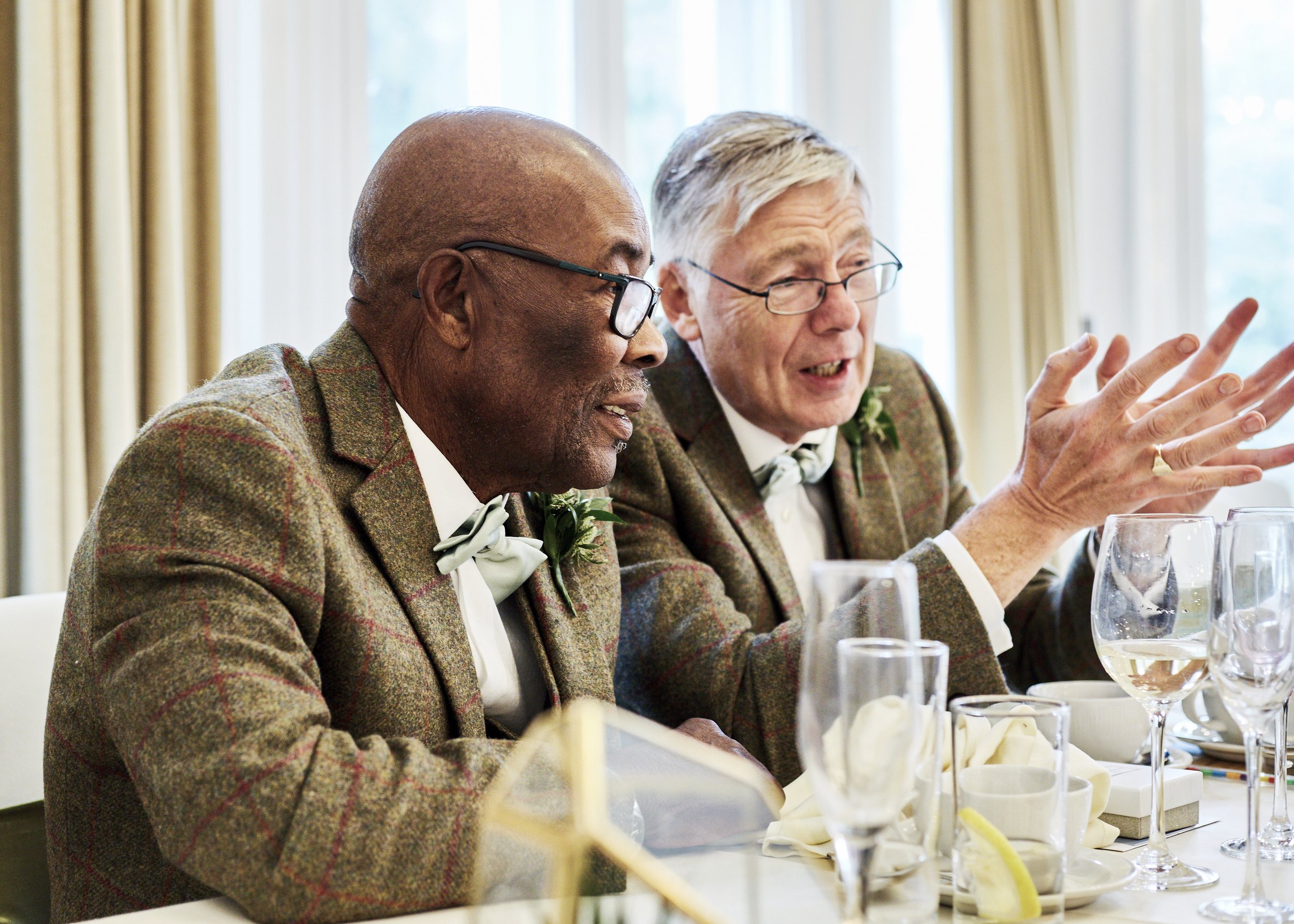 Two elderly men in tweed jackets and glasses sitting at a table during a formal meal, engaged in conversation. The table is set with wine glasses, pitchers, and napkins, with large windows and curtains in the background.