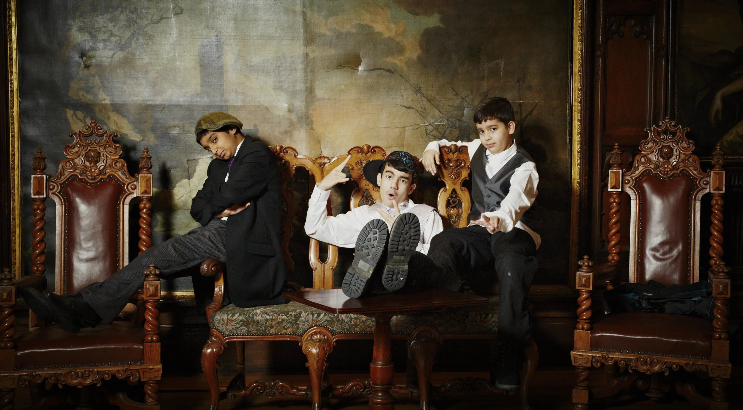 Four young boys dressed in vintage clothing posing on ornate chairs in front of a painted landscape backdrop with ships and clouds.