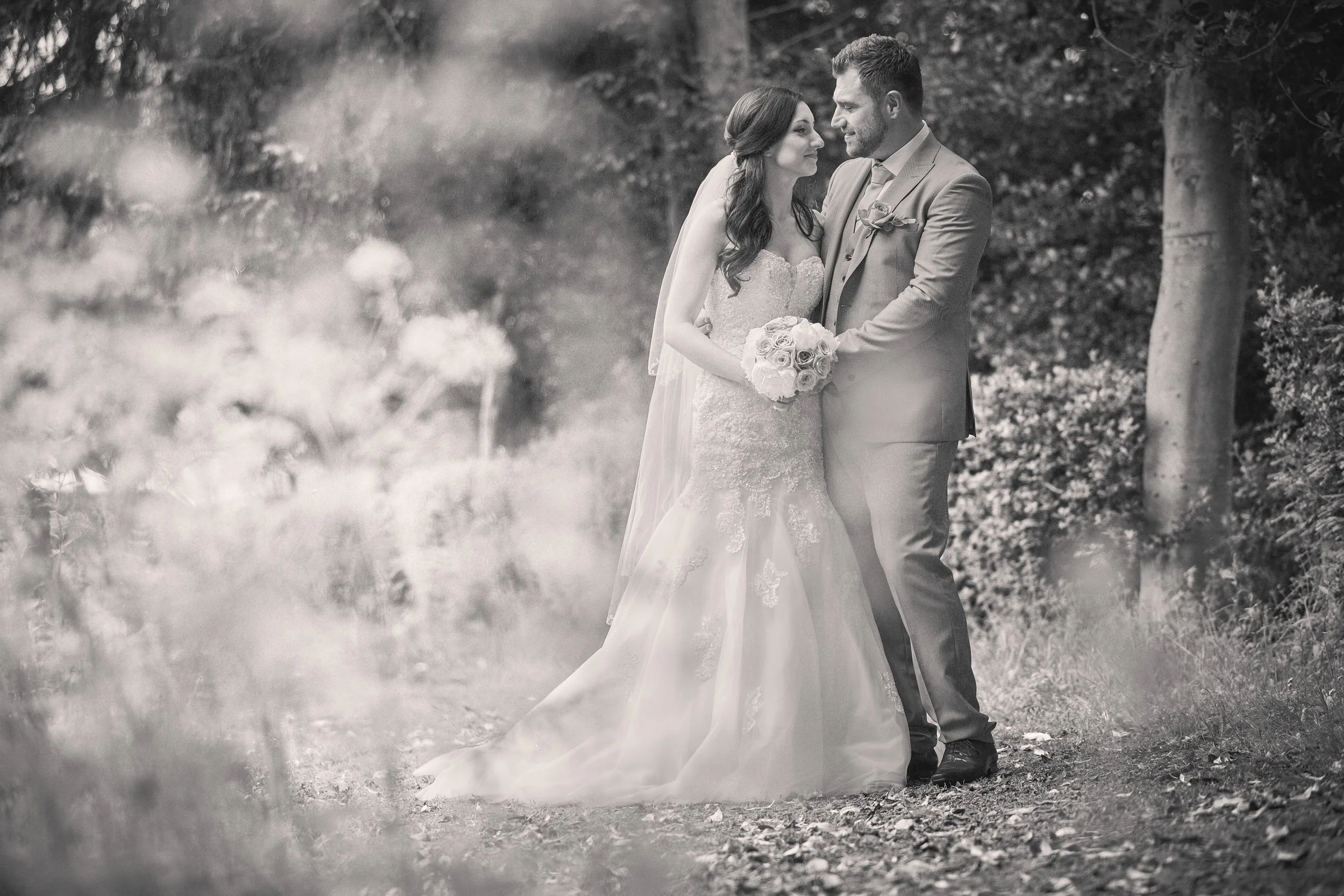 A bride and groom stand close together outdoors surrounded by trees, holding hands and looking into each other's eyes.