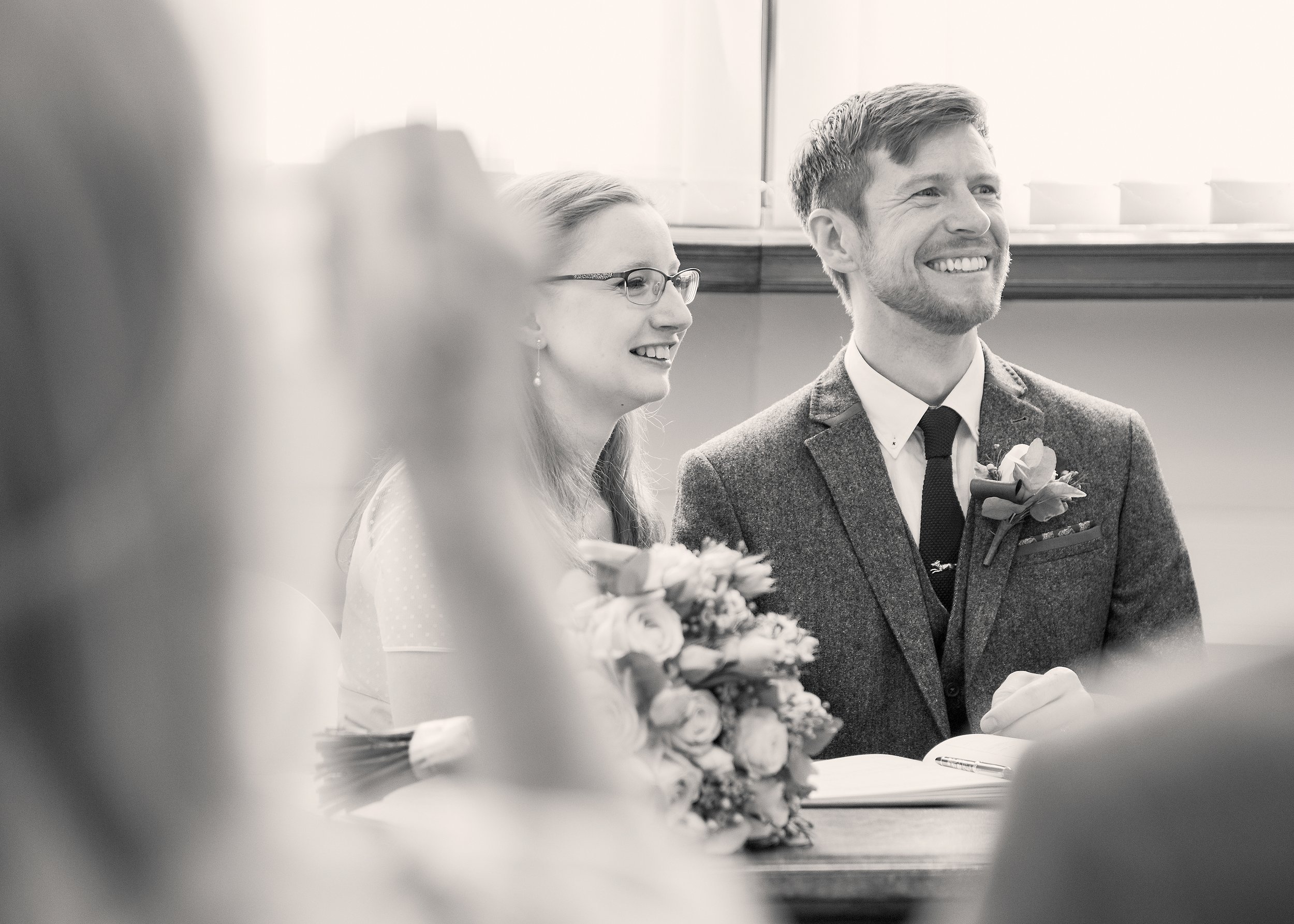 Black and white photo of a man and a woman smiling during a formal event, with women holding bouquets of flowers in the foreground.