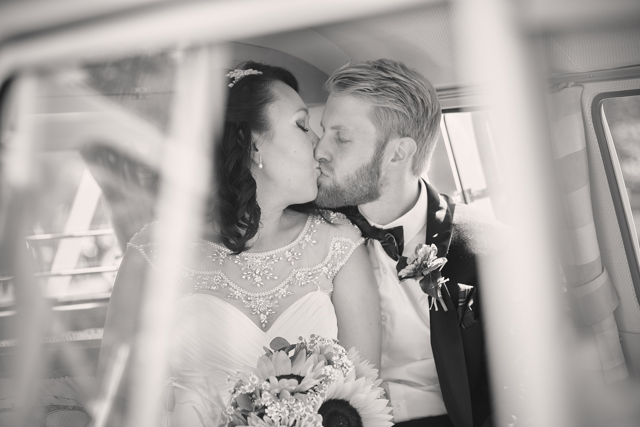 A bride and groom sharing a kiss inside a car on their wedding day, with the bride holding a bouquet of flowers.