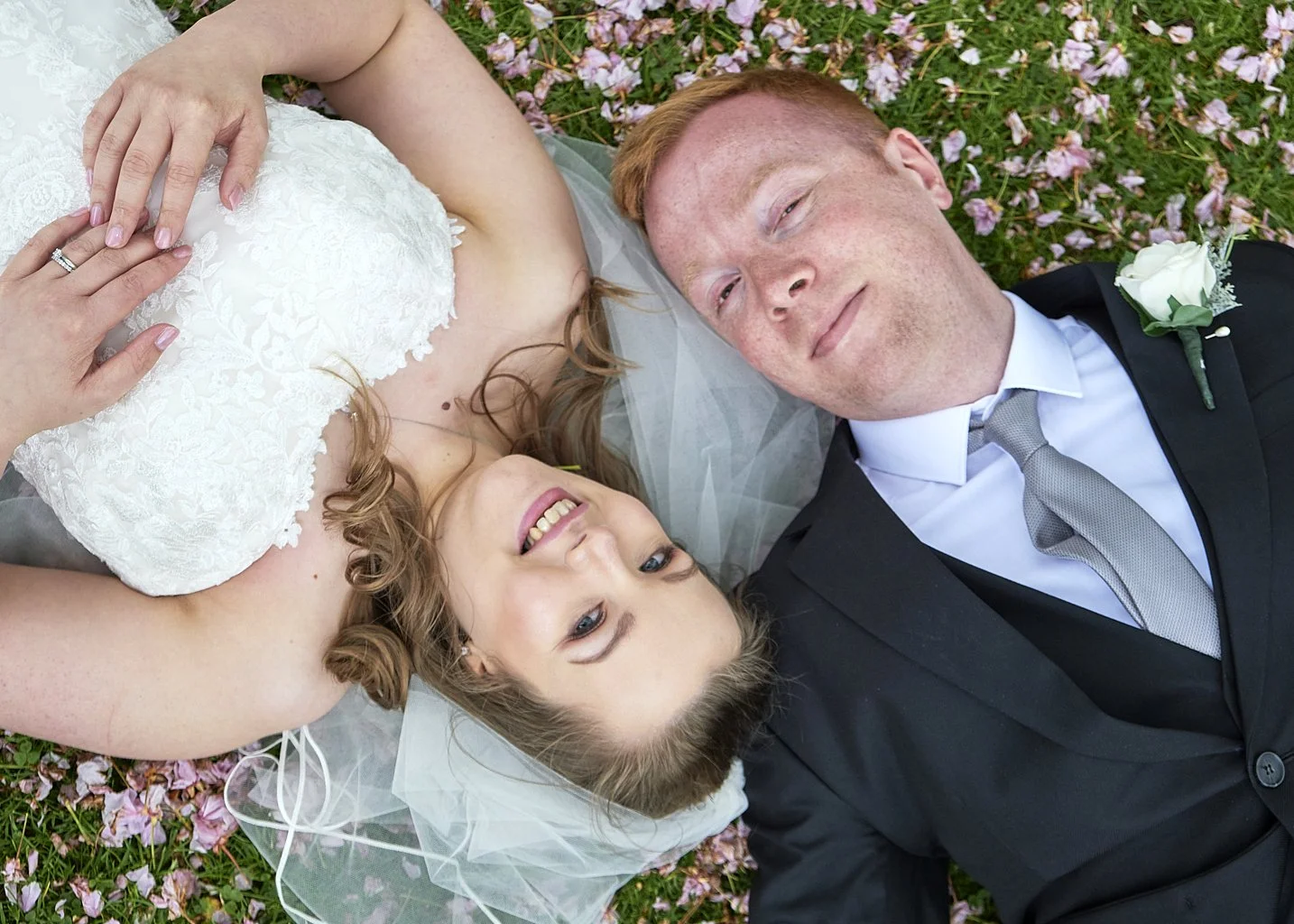 A newlywed couple lying on the grass covered with pink flower petals. The bride is wearing a white lace wedding dress and the groom is in a black suit with a gray tie and a white boutonniere. They are smiling and looking up at the camera.