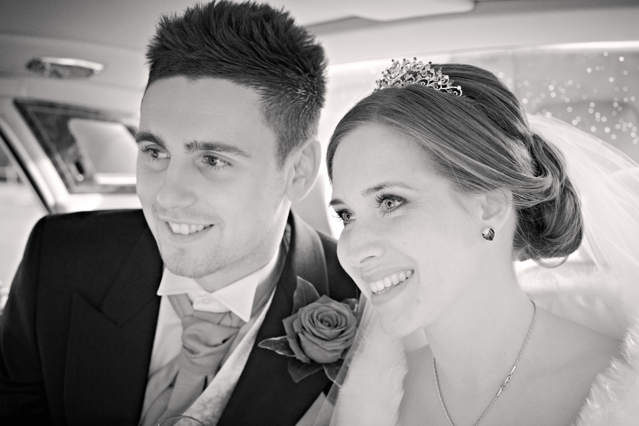 Black and white photograph of a smiling bride and groom, close-up on their faces, inside a vehicle, possibly at their wedding.