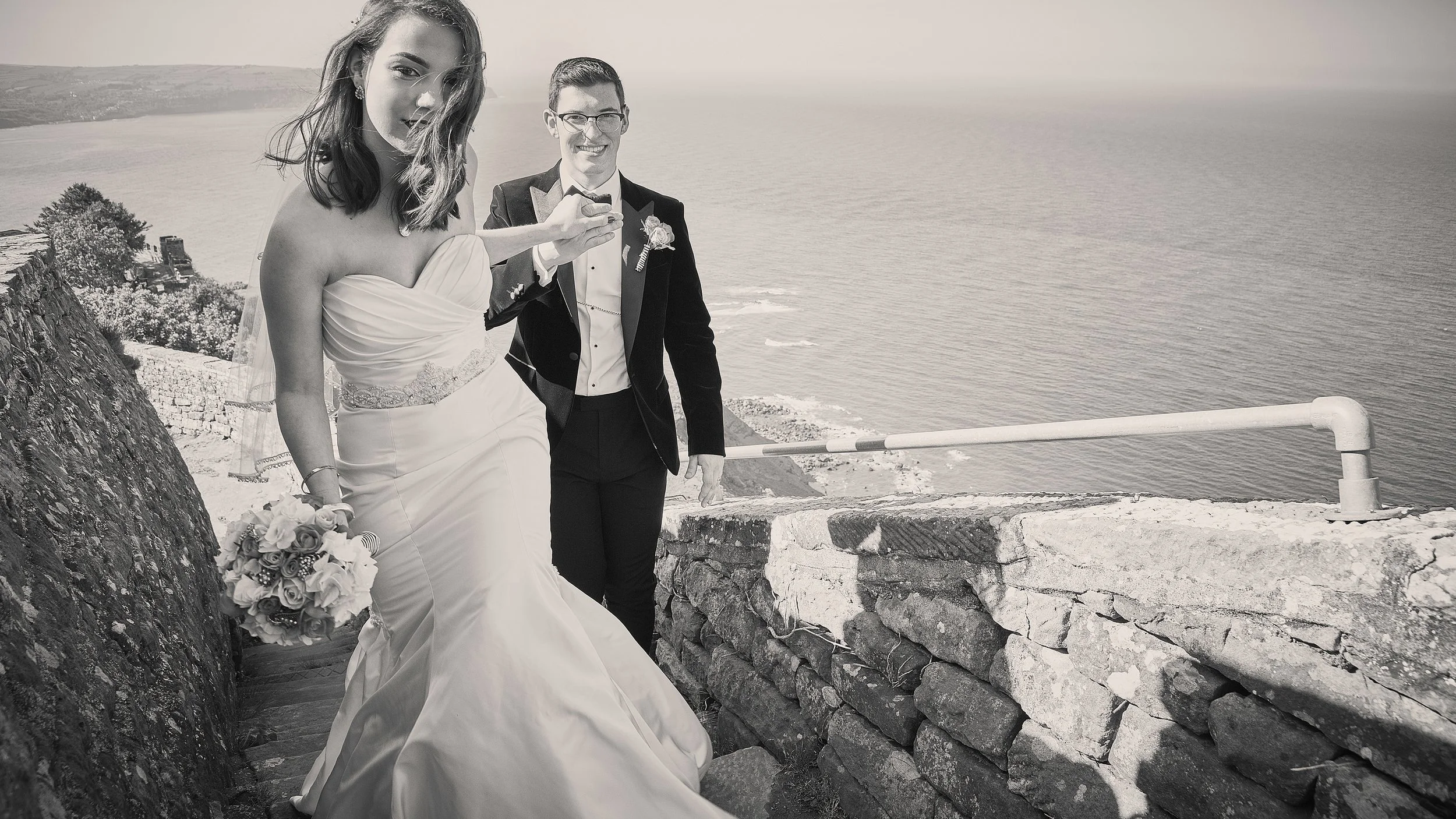A bride in a strapless wedding gown holding a bouquet, and a groom in a tuxedo, standing on a seaside walkway with a railing and stone wall, overlooking the ocean.