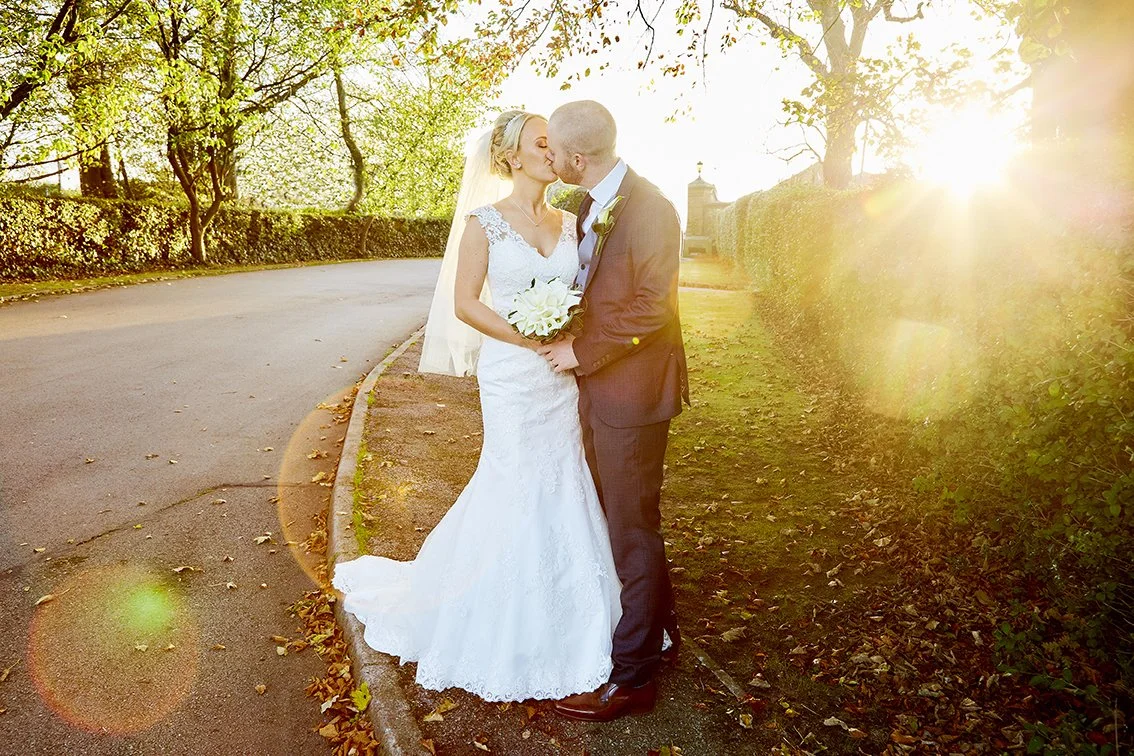A bride and groom sharing a kiss outdoors during sunset, with trees and a hedge in the background, on a paved path lined with fallen leaves.