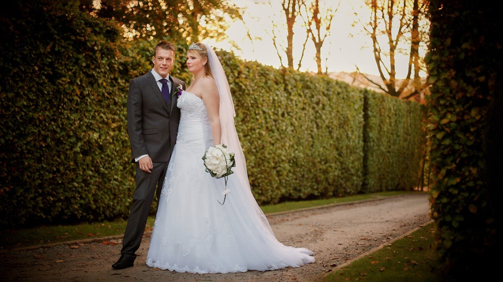A newlywed couple standing on a dirt pathway surrounded by tall, green hedges and trees with fall foliage at sunset. The bride wears a white strapless wedding gown and a veil, holding a bouquet of white flowers. The groom wears a dark suit with a pur