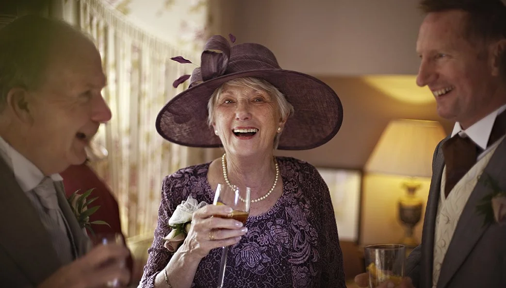 Three elderly people at a celebration, showing a woman with a large purple hat and purple dress smiling and holding a glass of wine, flanked by two men in suits, all appearing joyful.